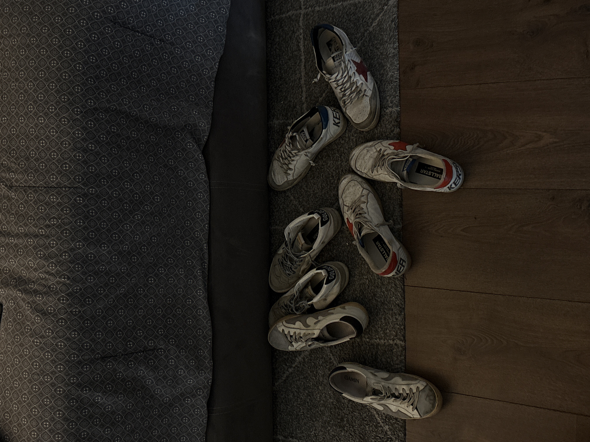 The scene depicts a collection of white, worn sneakers scattered on a gray rug that partially covers a dark wooden floor. The rug and shoes are positioned next to a bed or couch upholstered with a dark gray patterned fabric. There are no people visible in the image. The location is not possible to determine based on visual cues, as the setting could be in many places; therefore, San Bernardino Tlaxcalancingo, Mexico, cannot be confirmed or denied. The focus is on the arrangement of the shoes and the textures of the surrounding surfaces.