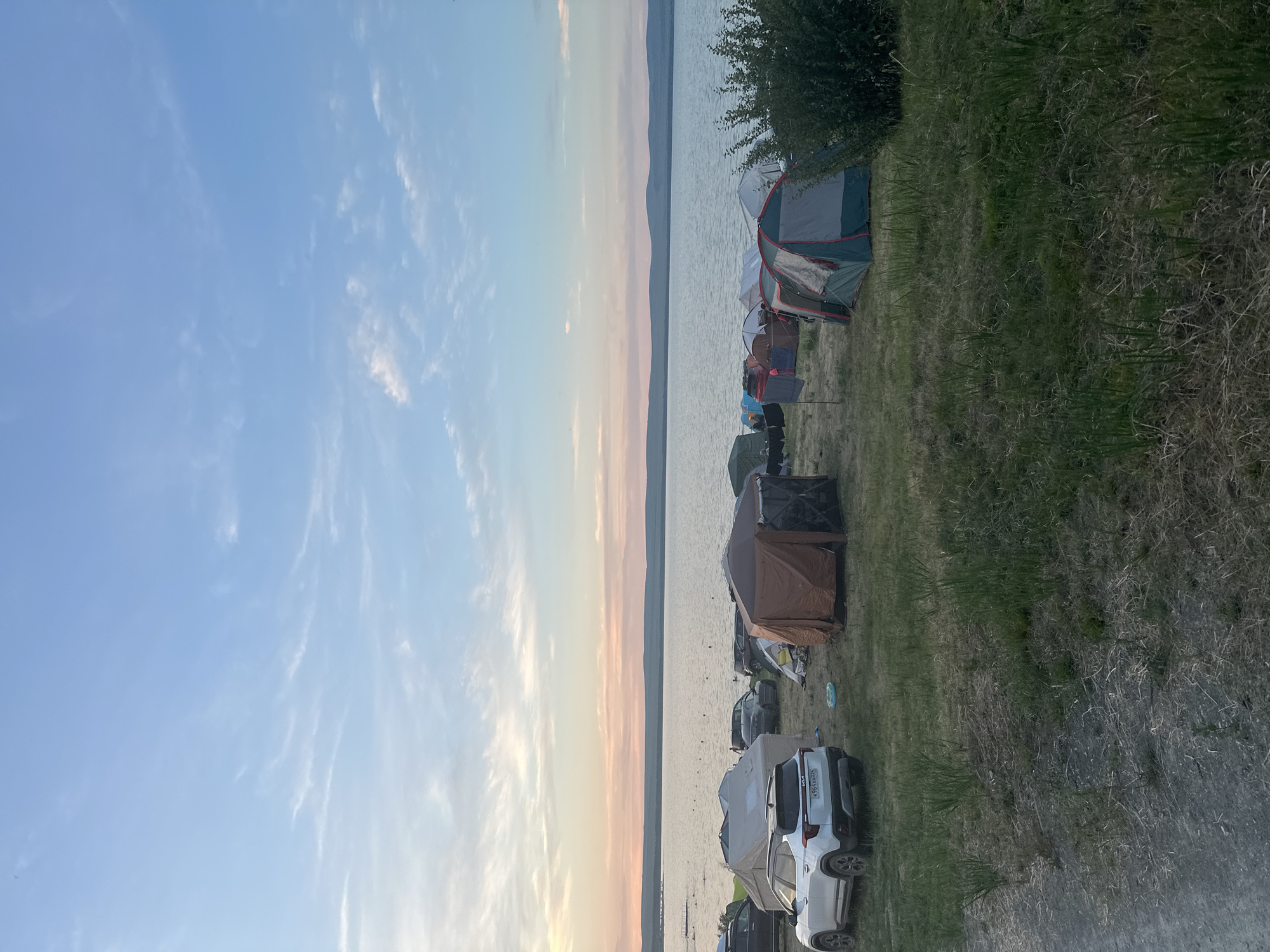 The image depicts a campground scene on the shore of a large body of water, likely a lake, under a sky with scattered clouds and the soft hues of either sunrise or sunset. Several tents are set up on the grassy terrain, varying in color and size, including a prominent tan and brown multi-room tent and a green and grey dome tent. Various vehicles, including a white Kia SUV and other cars, are parked among the tents. In the background, the calm water stretches to the horizon, with a distant, undulating shoreline. There are no visible people in the immediate foreground or mid-ground, though there might be figures in the water in the distance. The overall environment is that of an outdoor recreation area.