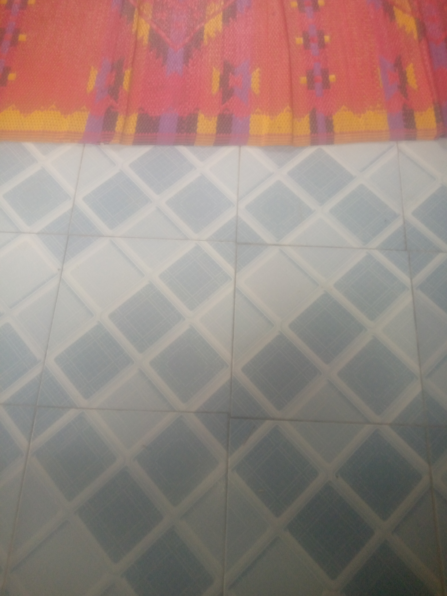 The image displays a close-up view of a tiled floor with a patterned textile draped across the top portion. The floor is covered in light blue tiles arranged in a diamond pattern, with white grout lines forming a grid. The textile, which appears to be a rug or curtain, features a vibrant geometric design in shades of red, orange, yellow, and purple. The lighting is soft, creating a somewhat diffused effect on both the textile and the floor. The image is taken from a straight-on perspective, with no people or identifiable objects other than the floor and the textile. The setting appears to be indoors, possibly a room or hallway. The location is not specified beyond "Unknown, Unknown."