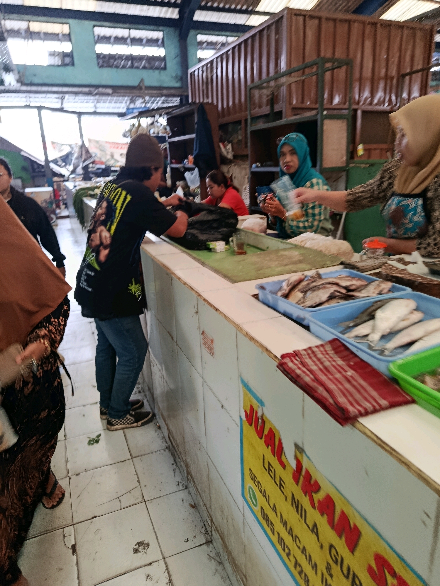 This is a photograph taken inside a market in Bululawang, Indonesia. The market appears to be a fish market, as evidenced by the trays of fish displayed on a counter. Several people are present in the scene. In the foreground, a person wearing a black t-shirt with a graphic on the back and blue jeans is standing at the counter, interacting with the market stall. To the left, another person wearing a brown hijab and a patterned skirt is partially visible. To the right, a person in a light-colored hijab is handing something to another person, possibly a customer. The market stall itself is made of white tiled material and has a yellow sign with red lettering that reads "JUAL IKAN" (Fish for Sale) and lists types of fish and a phone number. The environment seems to be an open-air market with a corrugated metal roof. The overall impression is of a bustling, working market environment.