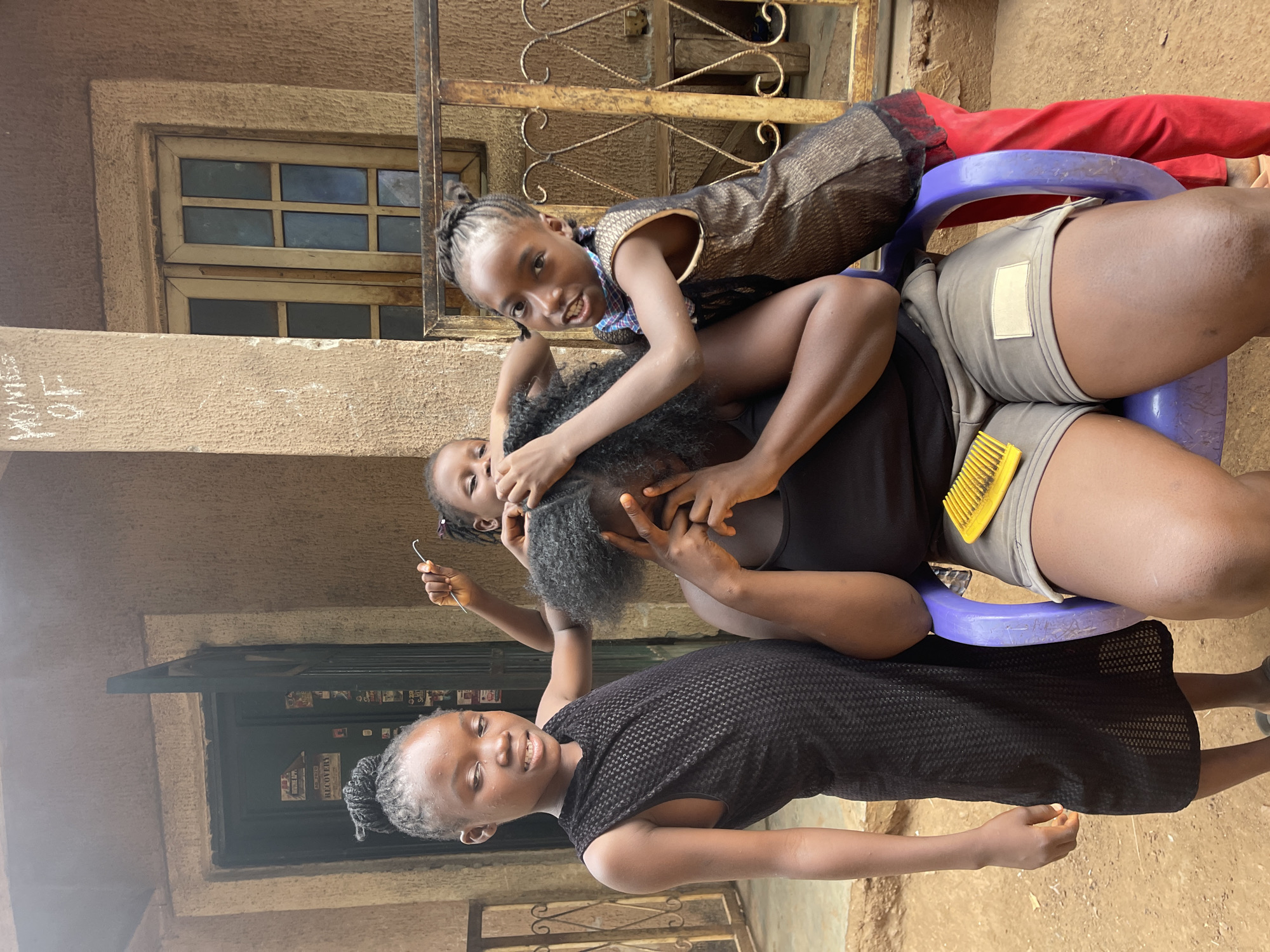 Four individuals are captured in an outdoor setting, likely in Akwanga, Nigeria, engaged in hair braiding. An adult, wearing a dark top and shorts, is seated on a purple plastic stool. Their dark, textured hair is being braided by a child who stands behind them. Another child, wearing a patterned top and shorts, is seated on the adult's lap, also seemingly involved with the hair and smiling towards the camera. A yellow comb rests on the adult's right thigh. A third child, wearing a black patterned dress, stands to the left, smiling towards the camera with their hair styled in braids.
The scene takes place outside a building with textured, light brown walls. A wooden window with multiple panes is visible in the background, along with a dark-colored door. A metal gate or decorative grill is also present. The ground is unpaved, appearing as dirt or dusty earth. The bright lighting suggests it is daytime with clear, sunny weather.
Faint, light-colored text, possibly "ADONES 07", is visible on the wall to the left of the window. On the dark door, a white sign reads "RECOVERY". The individuals appear relaxed and engaged in the activity.