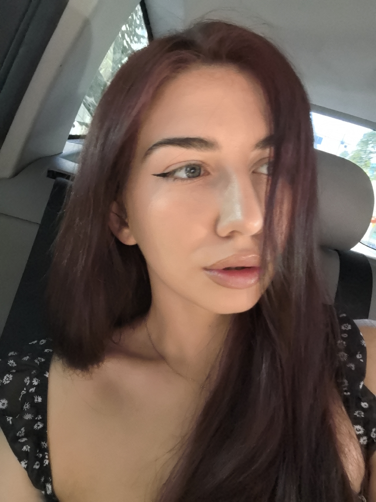 A head-and-shoulders portrait of a young woman is taken from inside a car in Stara Zagora, Bulgaria. She has dark reddish-brown, shoulder-length hair, parted on her left, with some strands covering part of her face. Her light-colored eyes feature prominent winged eyeliner, and her lips appear glossy. She wears a dark top with a small white floral pattern and a delicate necklace. Her gaze is directed away from the camera, towards the right, conveying a neutral or slightly contemplative expression. The interior of the car, including a headrest and window frames, is visible around her. Through the car windows, blurred greenery suggests an outdoor, daytime setting, indicating fair weather. There is no visible text.