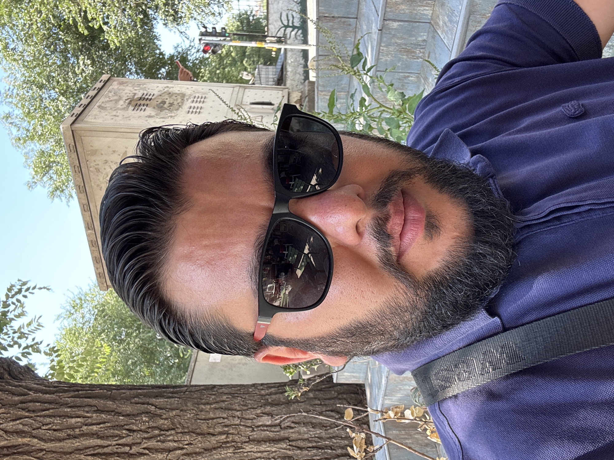The image is a close-up, waist-up selfie of a man with a beard and sunglasses, taken outdoors in Gungoren, Turkey. He is wearing a dark purple polo shirt and a dark strap is visible across his chest. In the background, a large tree trunk is on the left, with green foliage and a light-colored utility box behind it. A traffic light with a red signal is visible further in the background, suggesting an urban environment. The man's face is slightly turned, and the sunglasses reflect some of the surrounding scene, including what appears to be outdoor seating. The overall setting appears to be a sunny day.