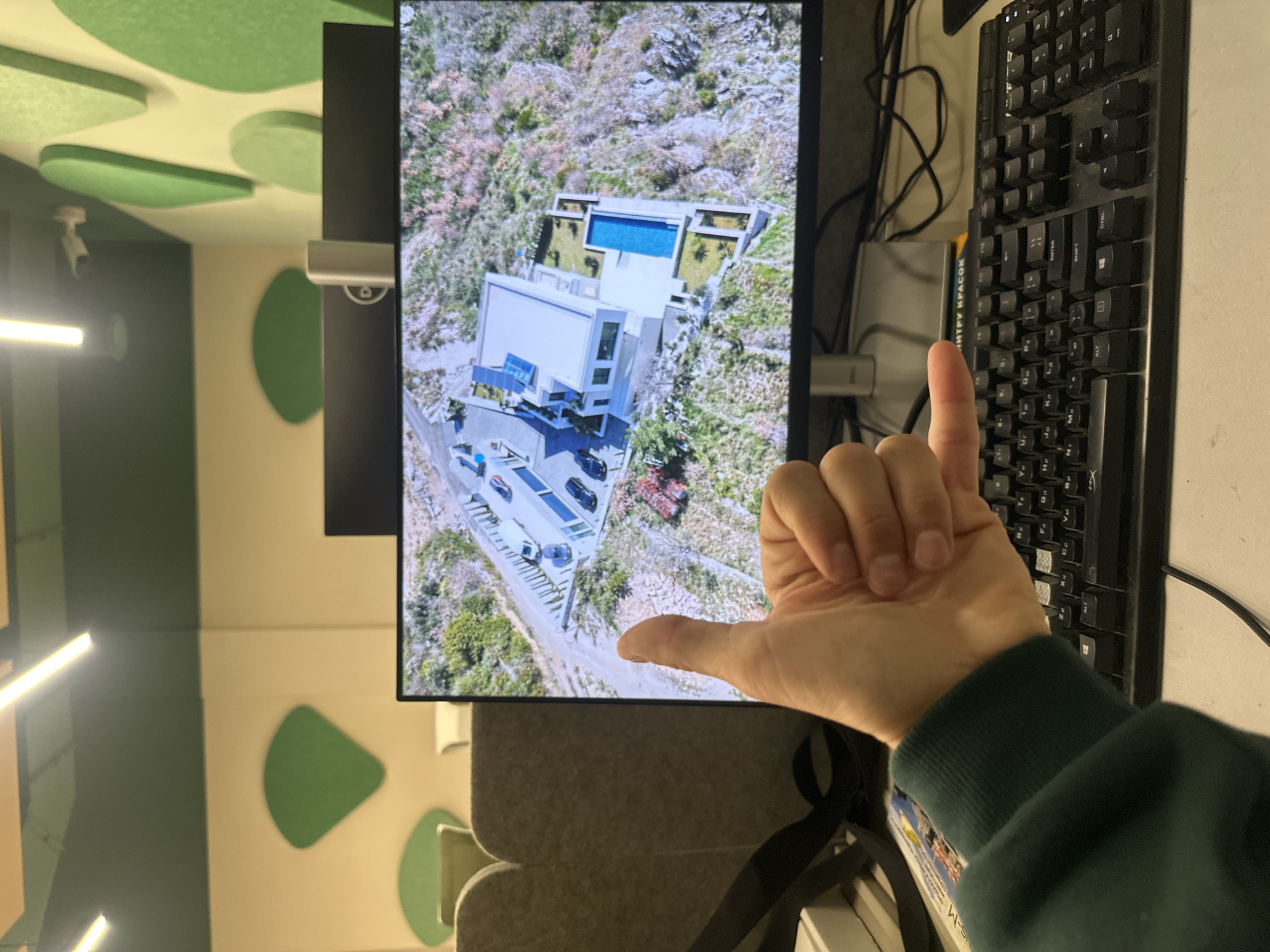 The image presents a screen displaying an aerial view of a modern house with a pool, situated within a landscape covered with trees and shrubs. The location is likely in Budva, Montenegro, as evidenced by the architectural style of the house and the terrain. A hand is making a "hang loose" gesture in front of the screen, and a black computer keyboard is positioned to the right. The surrounding environment appears to be an indoor workspace with a green and beige color scheme.