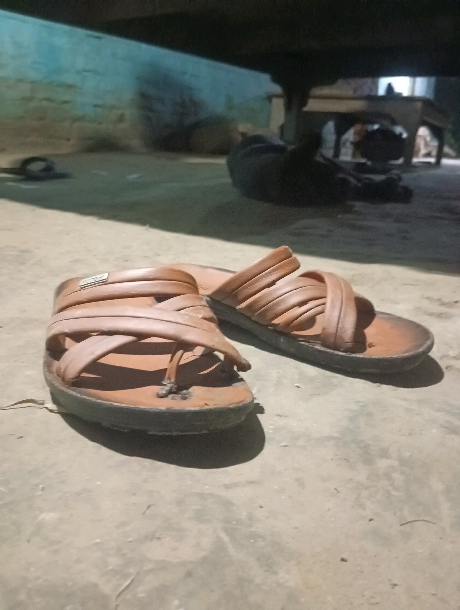 The image depicts an indoor scene, likely in Dakhin Parsa, India. In the foreground, a pair of brown leather sandals are visible, resting on a concrete floor. The sandals appear to be worn and are positioned close to each other. In the background, a dark, indistinct figure is lying down. The environment suggests a dimly lit, possibly unfinished or utilitarian space, with a wall visible in the far left and what appears to be furniture or structures further back. The lighting is dim, casting shadows.