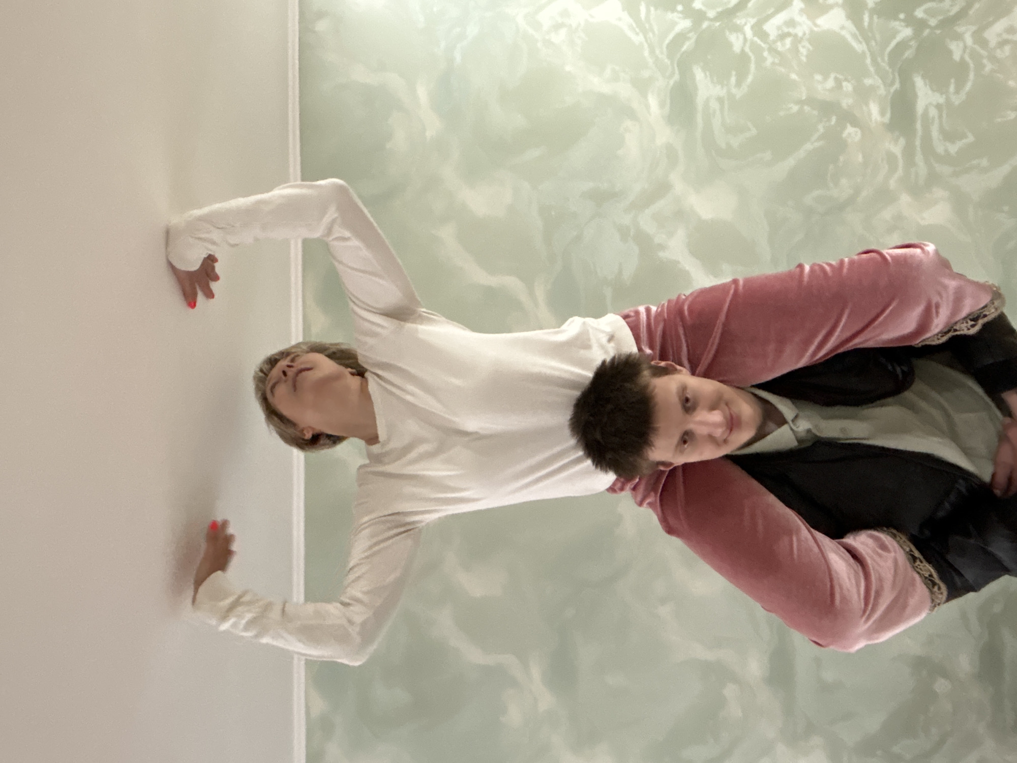 Two individuals are depicted in the image. One person is wearing a long-sleeved white top and has their hands pressed against a white wall. The other person is wearing a pink velvet jacket and appears to be positioned beneath the first person. The background features a wall with a marbled pattern. The exact location within the city of Chaikovsky, Russia, is not discernible from the image.