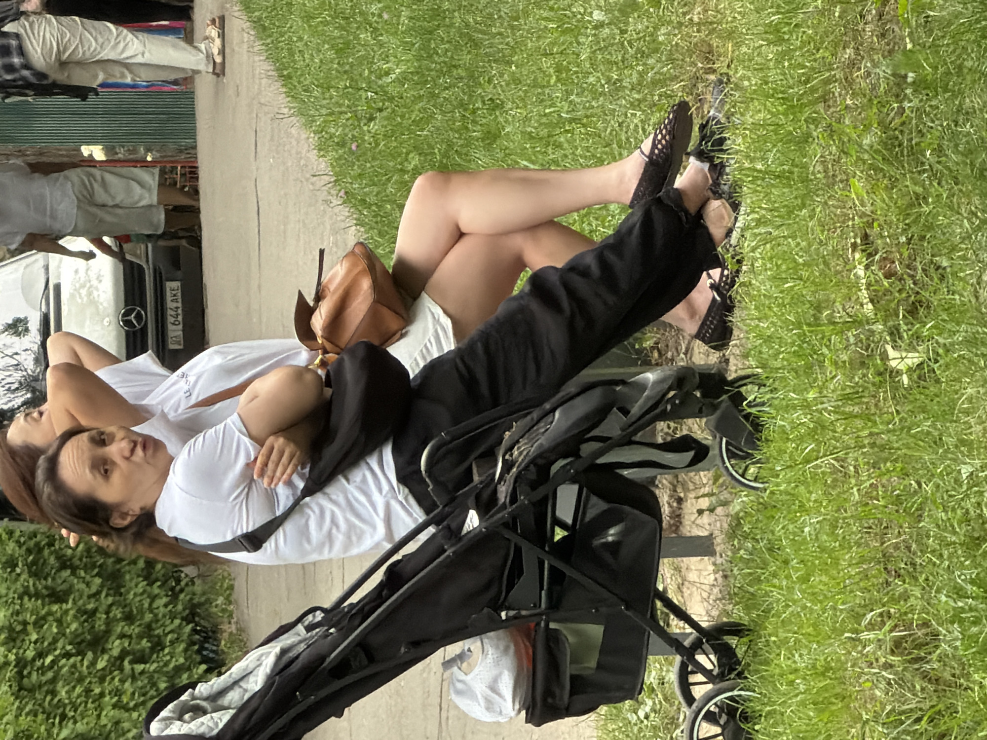 Two women are seated on a bench next to a stroller on a grassy area. The woman in the foreground, wearing a white t-shirt and black pants, looks towards the camera with her arms crossed. She has a black bag slung across her body and a brown purse on her lap. The woman behind her, wearing a white top and white skirt, is partially visible. The stroller is black and has a light-colored fabric lining. In the background, a silver van is parked, and a person in a plaid shirt and light-colored pants is walking away from the camera. A green metal fence is visible to the right. The scene appears to be outdoors in a park or public area.