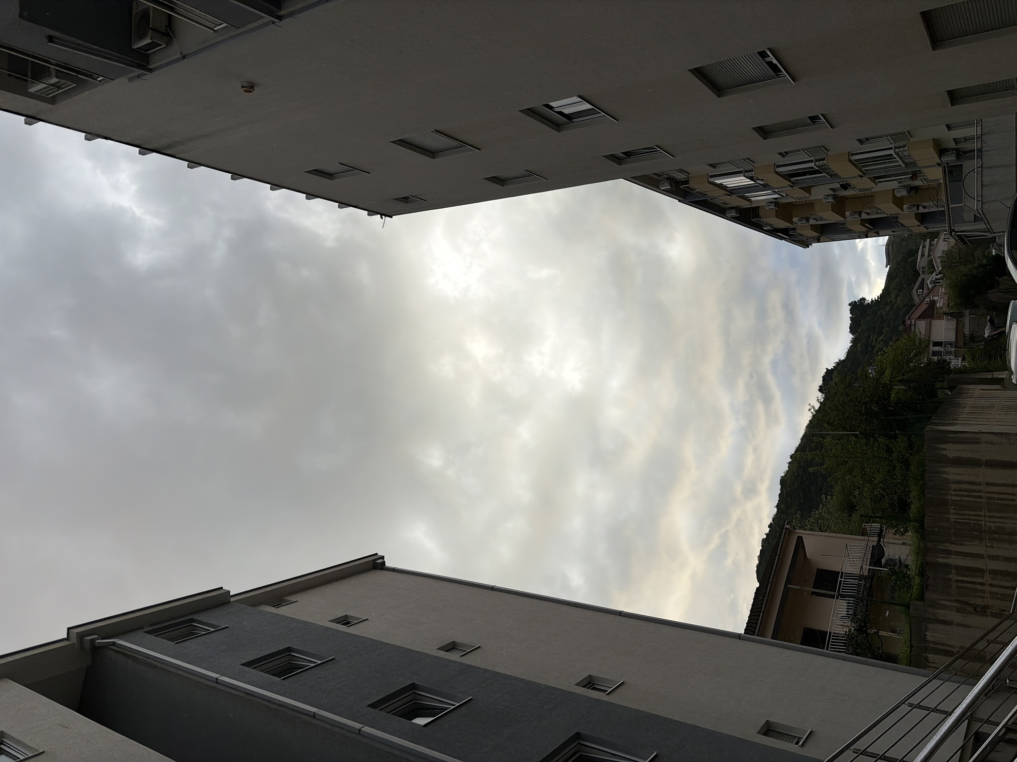 The image presents an upward-looking perspective, framed between two multi-story buildings, towards a cloudy sky and a distant, tree-covered hillside. The scene suggests an urban or suburban residential area, consistent with the hilly topography and coastal development found in Budva, Montenegro.
In the foreground, on the lower and left side of the frame, stands a modern building characterized by a dark grey lower section and a lighter, possibly beige or off-white, upper facade. It features multiple square windows, some with visible blinds or shutters. A metal railing, indicative of an external staircase or balcony, is partially visible in the bottom right corner.
Opposite this, bordering the top and right side of the image, is another substantial residential building. This structure appears to be an apartment complex, lighter in color, possibly white or pale yellow, with numerous windows, small balconies, and visible external air conditioning units. The underside of an overhang from this building is visible, revealing recessed features, possibly lights or vents.
The central and most expansive element is the sky, filled with dense, textured clouds. Brighter patches within the cloud cover suggest that sunlight is attempting to break through, indicating an overcast but not entirely dark daytime. On the far right, beyond the buildings, a lush, green hillside densely covered in trees and vegetation rises steeply. Glimpses of other residential structures, including houses with visible roofs and white exteriors, are nestled into the hillside, emphasizing the compact integration of development with the natural landscape.
No people are visible, and no specific activity is taking place; the image captures a static architectural and natural landscape. There is no discernible text within the frame. The overall impression is one of urban density combined with natural elements under a cloudy, daytime sky.