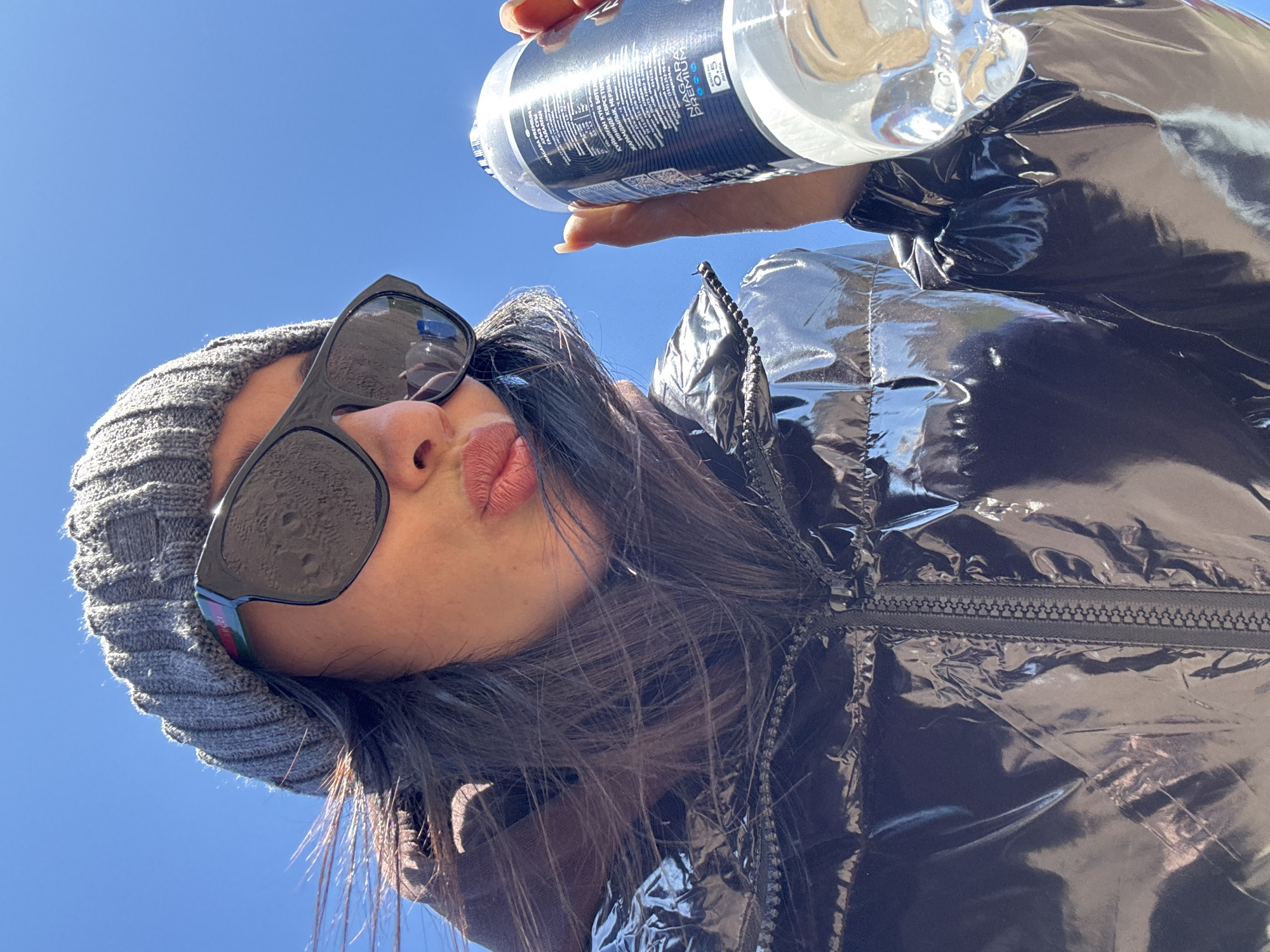 The image shows a woman from a low-angle perspective, looking down at the camera. She wears a gray beanie, sunglasses, and a shiny, dark puffer jacket. Her lips are puckered. She holds a bottle of water overhead, its label partially visible, with the text "NIAGARA PREMIUM," and Cyrillic characters indicating bottling and expiration details. The background is a clear blue sky. The text in the water bottle is partially illegible but includes the number "454136." The partial word "KҮЙЫЛҒАН" and number "0,5" are visible. The city and country are not directly visible within the image, but are referenced in the instructions.