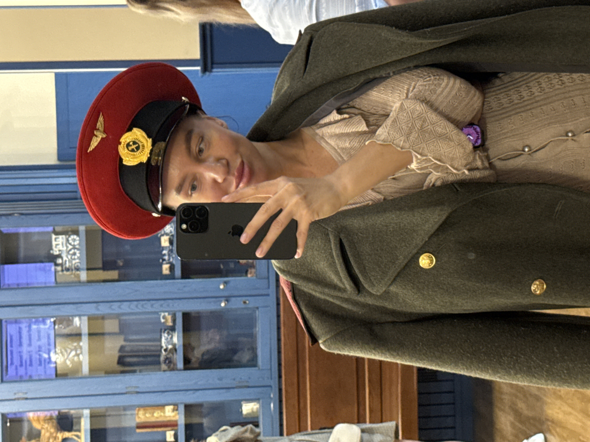 A woman is taking a selfie in front of a blue display case with artifacts, possibly in a museum or antique shop. She is wearing a red military-style hat with a gold emblem, a tan-colored blouse, and a heavy, dark green military coat. The phone she is using is dark gray or black. In the background, there's a wooden cabinet, and the location is likely in Yekaterinburg, Russia, due to the presence of Soviet-era memorabilia.