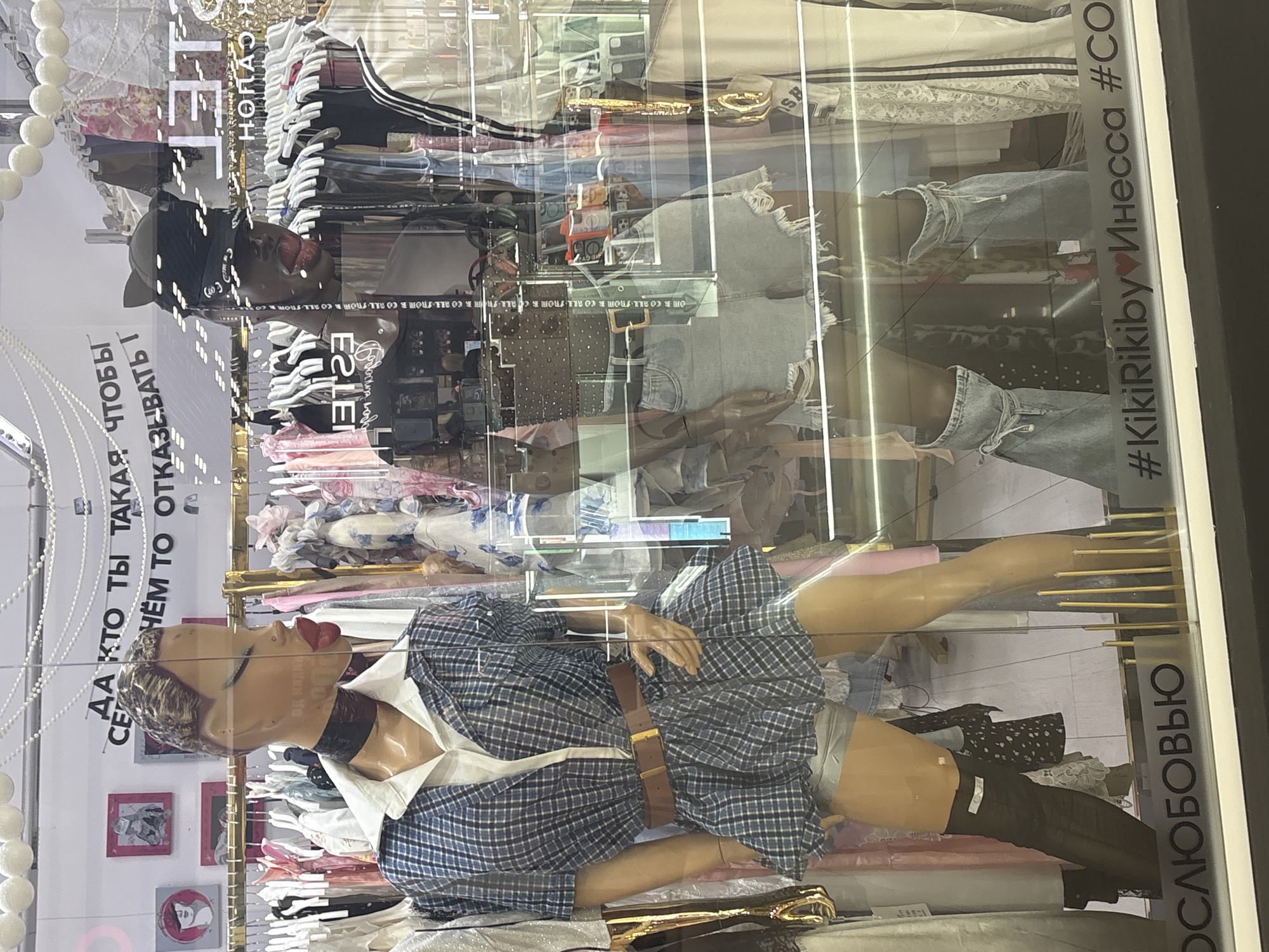 A store window displays mannequins dressed in fashionable clothing. One mannequin wears a blue plaid dress with a brown belt and black thigh-high stockings, while another mannequin is dressed in distressed denim shorts. The window also features lettering that reads "#KikiRikiby Инесса #CO", "ОСЛЮБОВЬЮ", and Russian text. The store is located in Tri Potoka, Russia.