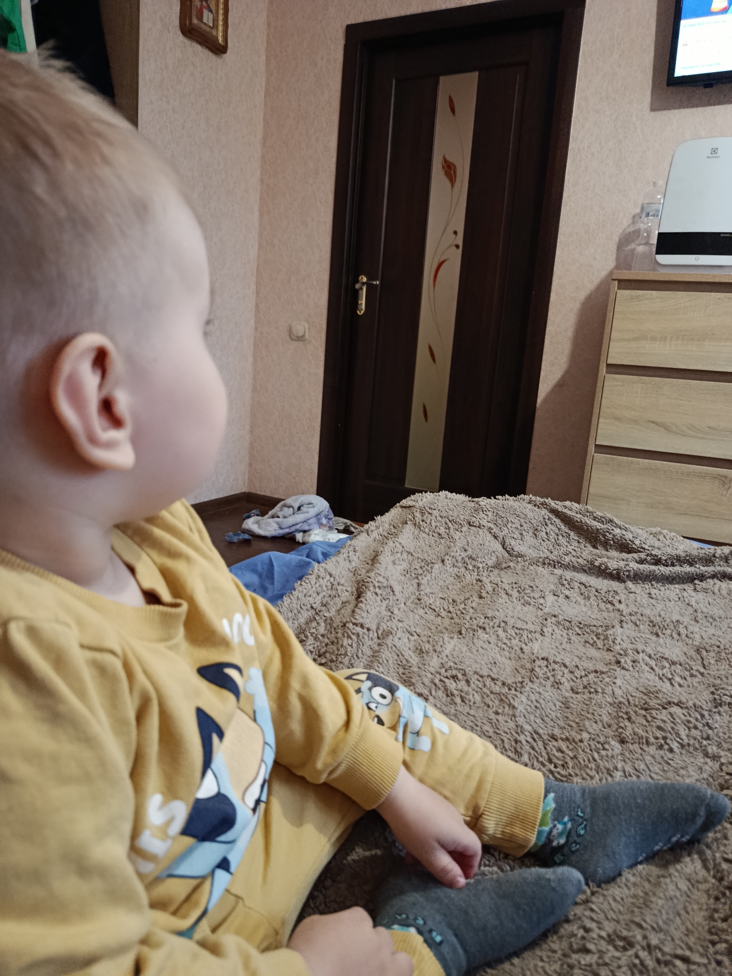This image, taken in the city of Unknown, Unknown, displays an indoor domestic scene. A young child, appearing to be a toddler, with light blonde hair, is seated on a textured, light brown blanket, facing towards the viewer's right. The child is wearing a mustard yellow long-sleeved shirt and matching pants, both featuring a multi-colored cartoon graphic. Dark grey socks with light blue patterns cover the child's feet.
The background features a light-colored, textured wall. On the left side, a small framed picture is mounted on the wall. Below it, a dark brown interior door with decorative vertical panels, a central light-colored accent with a reddish leaf motif, and a golden handle is visible. A white electrical outlet or switch is located on the wall near the door. On the right side of the background, a light wood-toned dresser with multiple drawers is present. On top of the dresser, a white and black Electrolux air purifier or humidifier stands next to a clear plastic water bottle. Above the dresser, a flat-screen television is mounted on the wall, displaying colorful content. The floor, visible beneath the door and dresser, is dark wood or laminate, and several crumpled textiles are present on it.