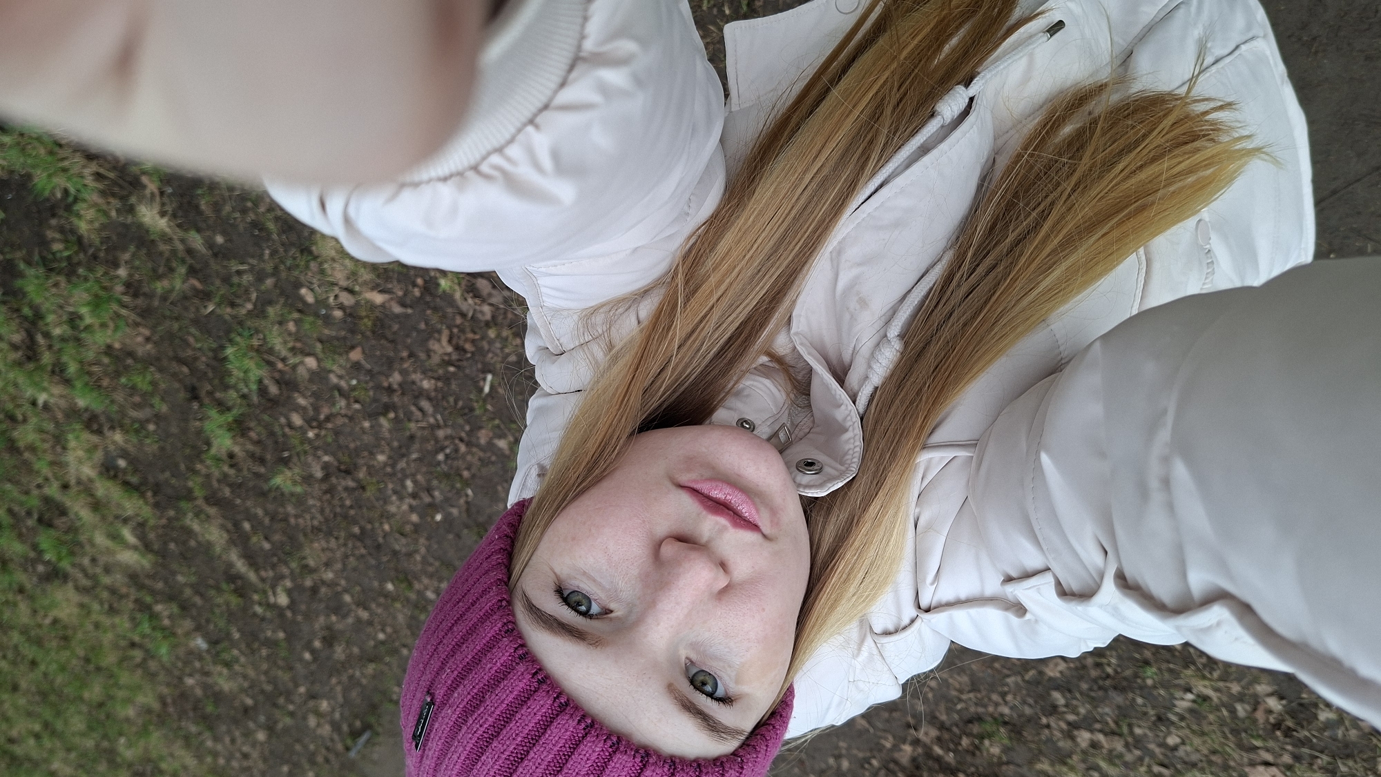 A woman is taking a selfie outdoors. She is wearing a pink knit hat and a light beige puffer jacket. Her long blonde hair is parted in the middle and falls down over her jacket. The background consists of dirt and sparse grass, suggesting a park or undeveloped area. The photograph was taken in the city of Chaikovsky, Russia.
