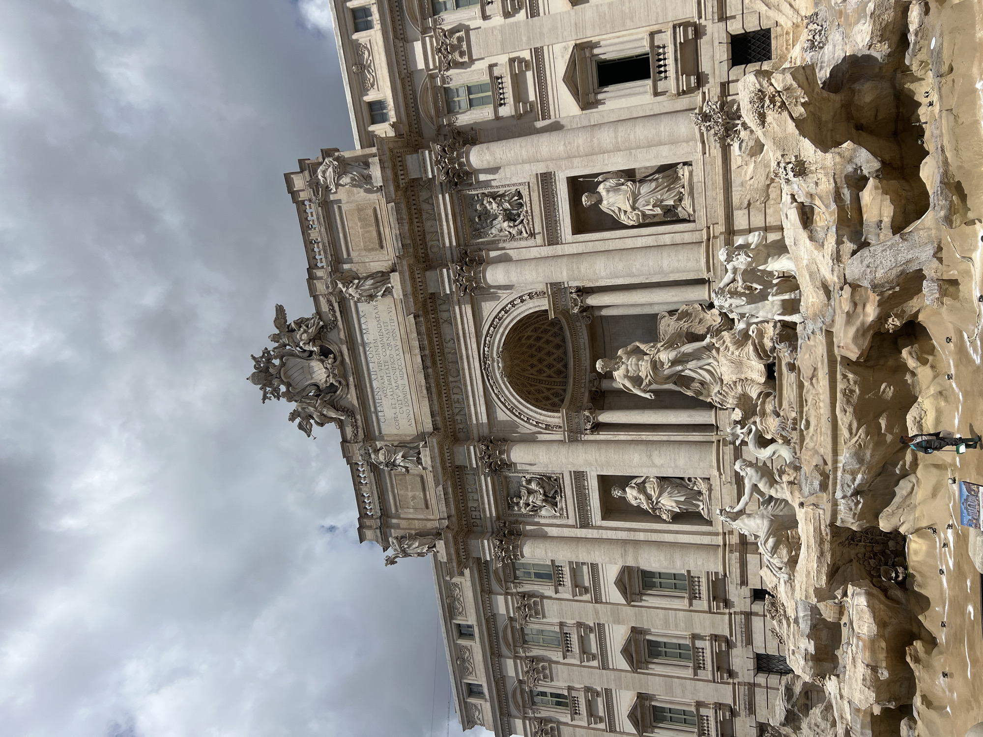 The image shows the Trevi Fountain in Rome, Italy, under a cloudy sky. The fountain's baroque architecture is on display, featuring statues of figures such as Oceanus riding a chariot pulled by sea horses. A person stands near the water's edge on the right, partially obscured by the rock structures of the fountain. A small sign or advertisement is visible near the person. The architecture of the Palazzo Poli behind the fountain can also be seen.