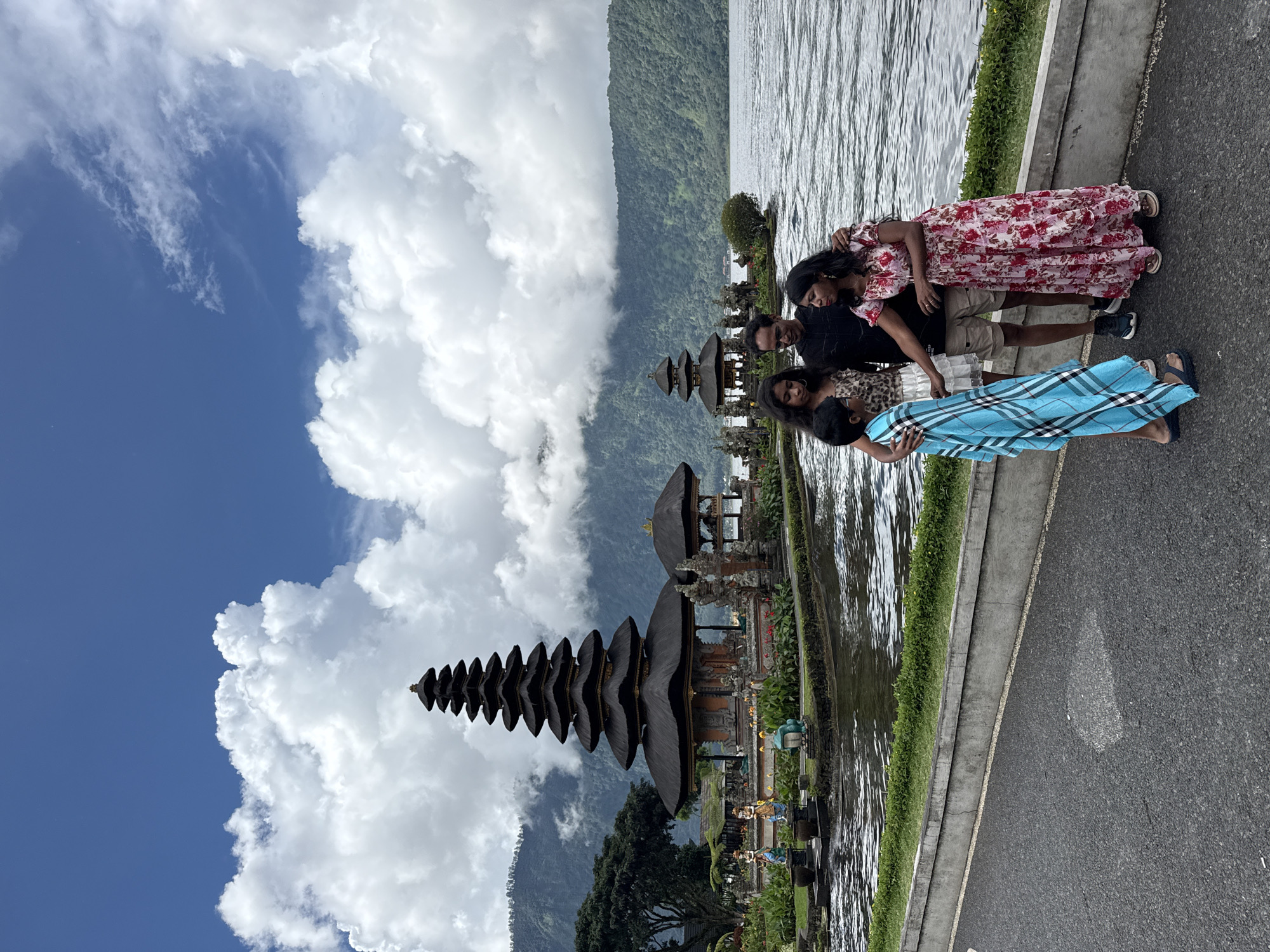 A medium shot shows a group of four people standing on the edge of a paved road, with a water feature and a temple structure behind them. The temple, with its multi-tiered roof, is reflected in the water. Behind the temple and water, a hillside is partially obscured by clouds. To the left, a blue sky is filled with large, white cumulus clouds. The photograph was taken in Baturiti, Indonesia.
The group includes one man and three women. The man is dressed in a dark shirt and khaki shorts, one woman is wearing a blue dress, another is wearing a floral patterned dress, and the third is wearing a patterned dress. They are standing close together and are facing towards the temple.