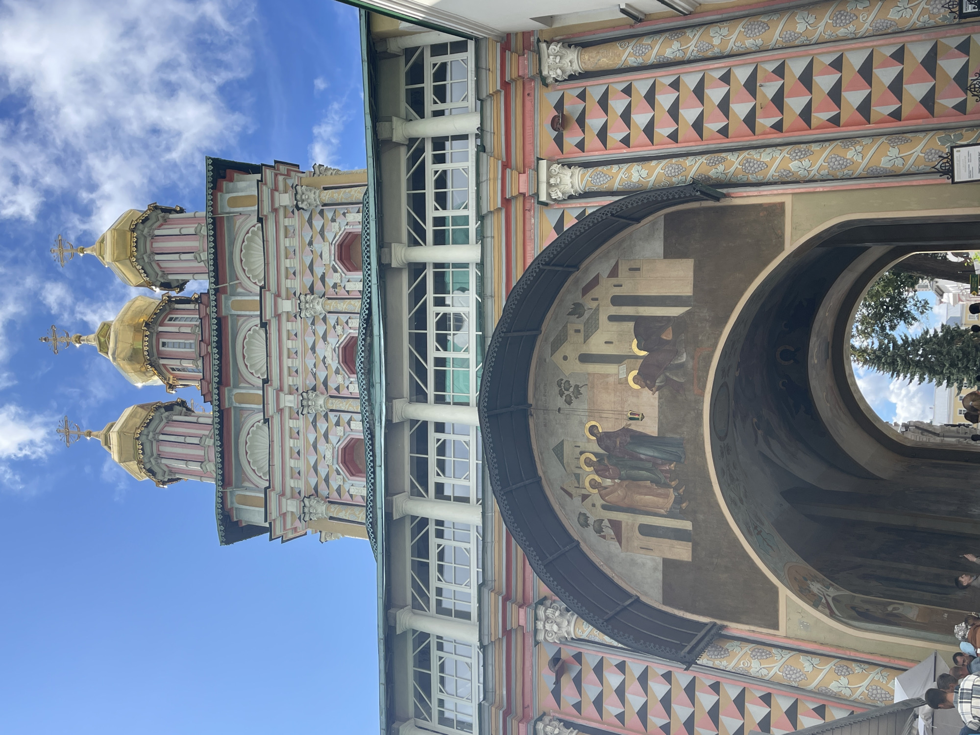 The image shows a low-angle view of the ornate Trinity Lavra of St. Sergius in Sergiev Posad, Russia. The architecture features multiple golden domes topped with crosses, painted in pink and white decorative patterns. Below the domes, a structure with arched windows and a large fresco depicting religious figures is visible. The fresco shows several individuals in robes, with halos, and a lamp hanging nearby. The lower part of the image reveals an arched passageway leading into the complex, with a tall evergreen tree visible in the distance. Several people are standing near the entrance to the passageway, some appearing to be tourists. The sky is blue with scattered white clouds. The building displays vibrant colors, including yellow, pink, and white, with decorative columns and geometric patterns.
