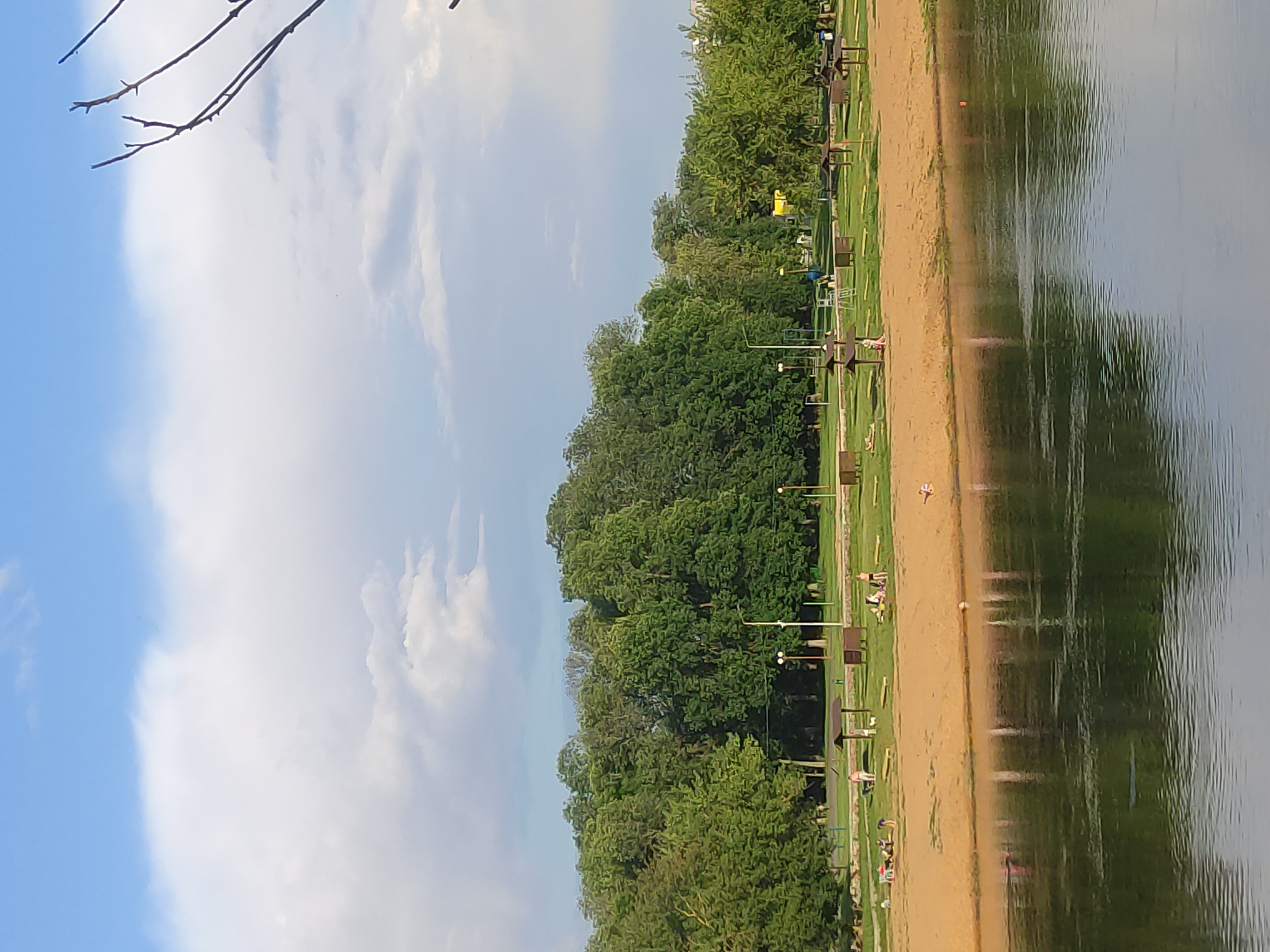 This is a wide-angle, eye-level shot of a park area with a sandy beach along a riverbank. The sky is partly cloudy with large white clouds against a blue background. In the foreground, the dark, rippling surface of a river reflects the trees and sky. Beyond the river is a sandy beach with several people scattered about, some sitting on towels or blankets and others standing. There are picnic tables and sheltered areas, some with dark brown roofs, positioned on the grass behind the beach. A line of dense green trees forms the backdrop to the park area. In the far distance, a few buildings are visible. The overall scene depicts a recreational area on a warm day. Based on the provided context, this location is in Mahilyow, Belarus.