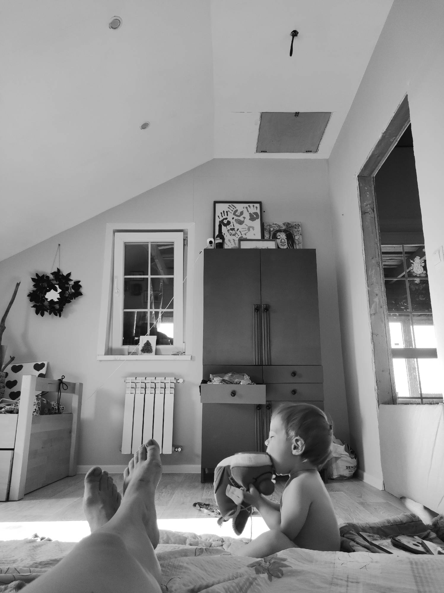A grayscale image captures a scene in what appears to be a room under renovation, possibly in a private residence. In the foreground, a pair of bare feet are visible atop a patterned blanket, suggesting someone is lying down. A child, shirtless, sits upright on the blanket, engaged with a stuffed toy, putting it to their mouth.
In the background, there is a combination of domestic elements and signs of construction. A dark wardrobe stands against the wall, adorned with various framed pictures on top. A window with multiple panes is present, fitted with a radiator beneath it. To the left, a wreath and other decorative items are placed on or near a small shelf.
Adjacent to the wardrobe, an unfinished doorway reveals exposed wooden framing, indicating ongoing renovation work. The ceiling has at least two recessed lights and an access panel, adding to the impression that the room is under construction or being remodeled.
There is no verifiable information within the image to confirm the city of Фоки, Russia, as the location. Therefore, the presence of Фоки cannot be determined from this picture alone.