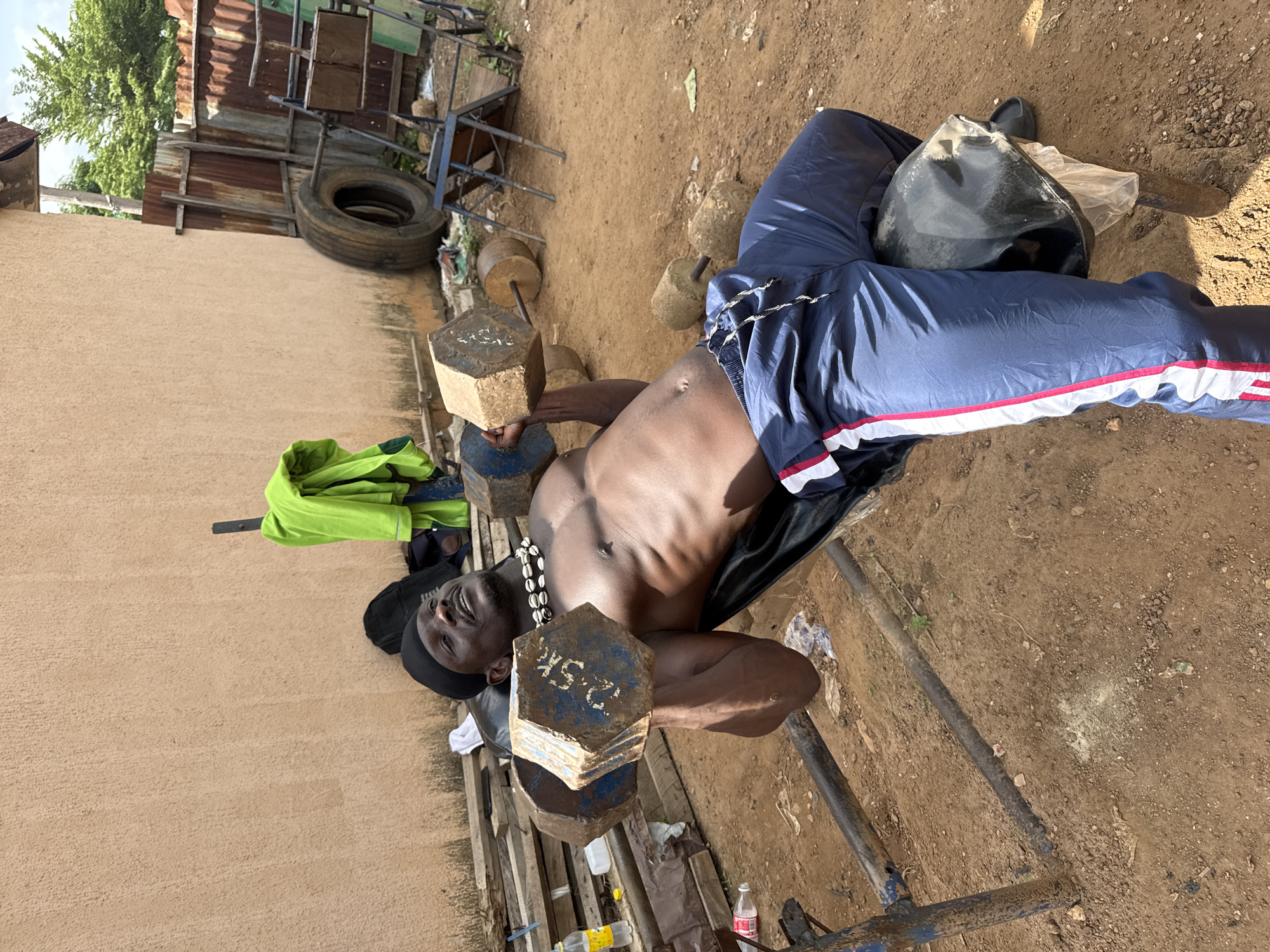 The image depicts an outdoor, makeshift gym scene in Akwanga, Nigeria, under bright daylight. The central subject is a muscular, shirtless man with dark skin, wearing a dark head covering and a beaded necklace. He is dressed in blue track pants with red and white stripes down the sides.
The man is lying on an improvised weight bench, constructed from metal pipes and wooden slats, performing dumbbell presses. He holds two large, hexagonal dumbbells, which appear to be homemade from a concrete-like material. One dumbbell, held closer to the viewer, has "2.5K" (likely indicating 2.5 kilograms) painted in white on its blue-painted surface. Another dumbbell held further back also shows faint numbers, possibly "25K".
The immediate surroundings consist of bare, dusty earth with scattered small rocks and debris, including plastic bottles and wrappers. In the background, a structure made of rusted corrugated metal sheets and wooden planks is visible, with an old car tire leaning against it. A bright green jacket or piece of clothing is draped over part of this structure. To the far left, a light-colored building wall extends, and lush green trees are seen against the sky. Sunlight casts strong shadows, indicating a bright, clear day, likely midday or afternoon. A black bag rests on the man's lap or near his legs. The scene highlights resourcefulness in creating exercise equipment and a training environment.
