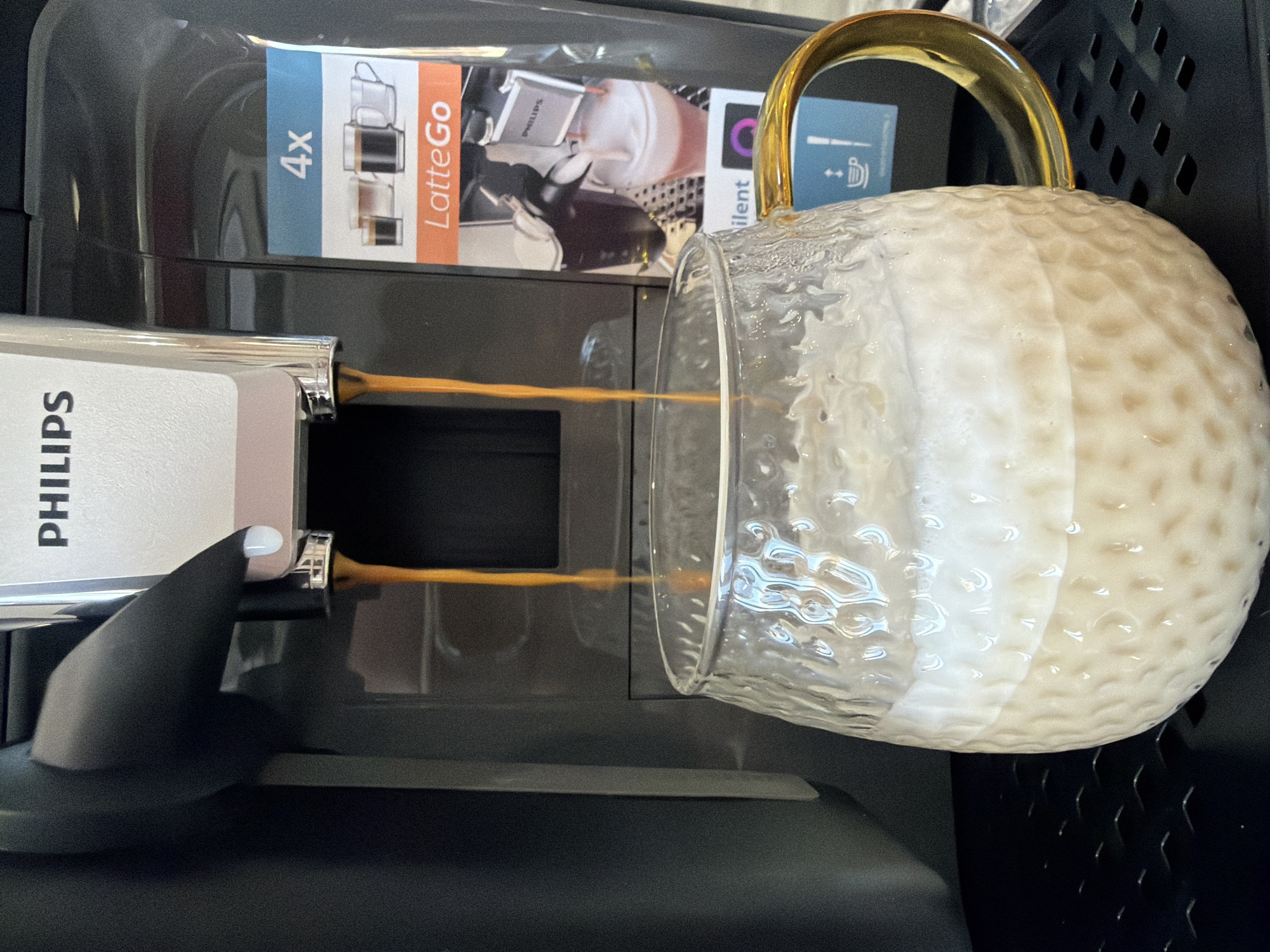 An automatic Philips coffee machine, dark grey and silver-grey, is actively dispensing two parallel streams of dark brown liquid into a clear, textured glass mug positioned beneath its spouts. The mug features an amber-gold colored handle on its right side and contains a layered beverage: a light brown, foamy base topped by a distinct, thick layer of white foam. The dispensed liquid is impacting this white foam. The machine's front panel displays the "PHILIPS" logo, and a vertical sticker on its right side features "4x" and "LatteGo" branding, alongside graphic representations of various coffee beverages. The coffee machine rests on a black, gridded drip tray. No individuals are visible in the frame. This scene is located in the city of Chaikovsky, Russia.