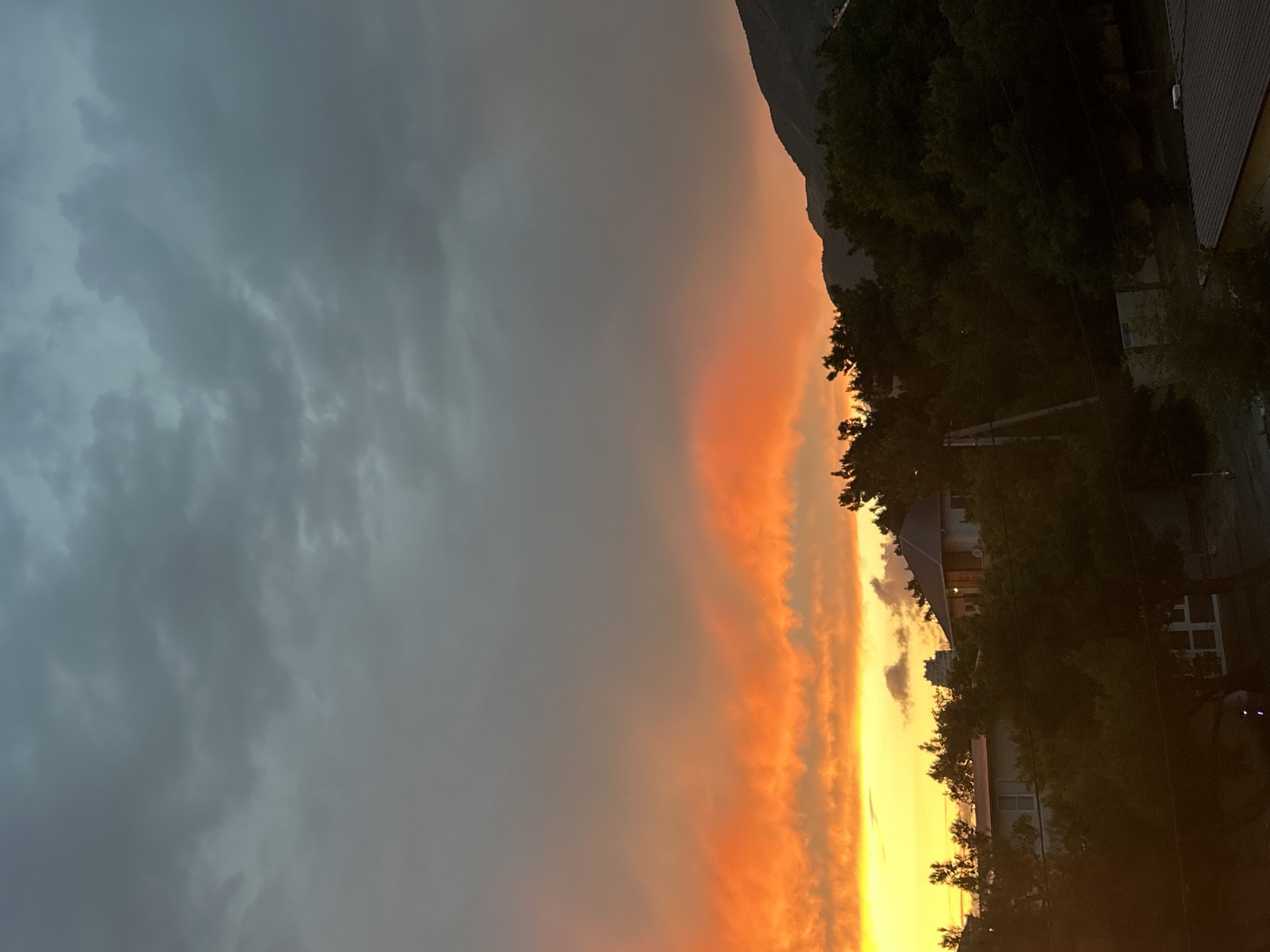 A sunset scene unfolds over Gelendzhik, Russia. The sky is dominated by dramatic clouds, with the lower portion ablaze in vibrant orange and yellow hues, suggesting the setting sun. Above this, the clouds transition to a darker, cooler gray. Silhouetted against the colorful sky are lush green trees and the rooftops of several houses. In the background, a dark mountain range rises, its peaks partially obscured by the trees and evening light. The overall impression is a tranquil, natural landscape at dusk, with human structures subtly integrated into the environment.