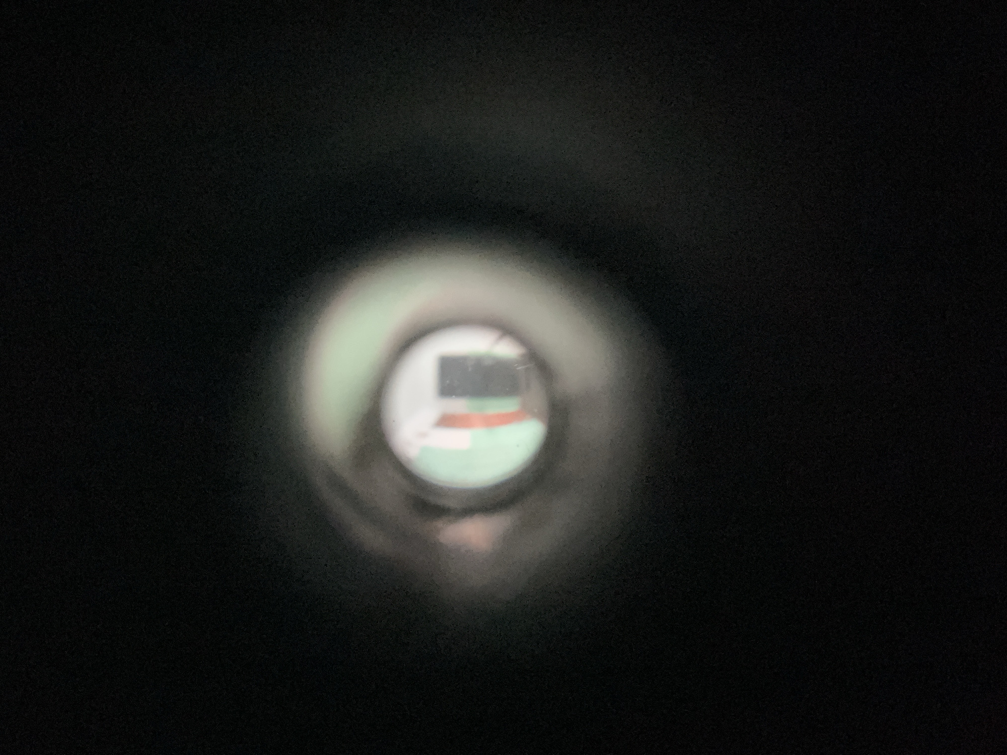 An eye is looking through a peephole. The peephole offers a blurry view of a hallway with a green and white striped wall. A dark rectangle, possibly a door or frame, is visible in the distance. The lighting suggests it is indoors. The location is Unknown, Unknown.