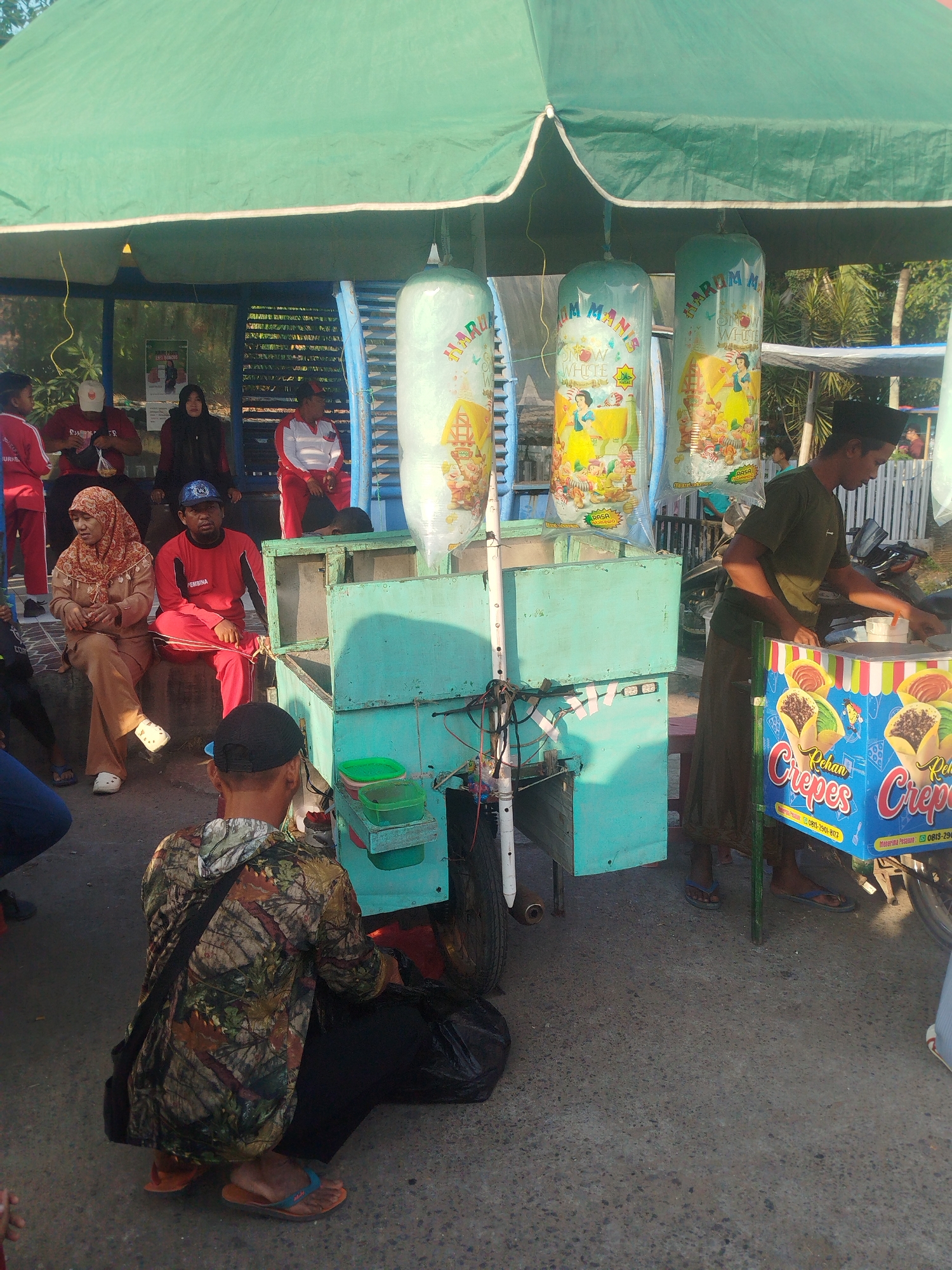 A group of people are gathered under a green awning, likely at a street food stall in Burneh, Indonesia. In the foreground, a man wearing a camouflage jacket and a black cap is squatting, facing away from the camera. To his right, a food stall is set up, featuring a turquoise wooden structure with various containers and equipment. A sign on the stall advertises "Rehan Crepes" with contact information. Behind the stall, a man wearing a green t-shirt and a sarong is attending to the food. In the background, several individuals are seated on a bench under the awning, including a woman in a hijab and a man in a red tracksuit. There are also three large, clear plastic bags filled with what appear to be beverages hanging from the awning, decorated with colorful images. The ground is paved with asphalt. The scene appears to be a typical street-side vending operation, possibly in a market or public area.