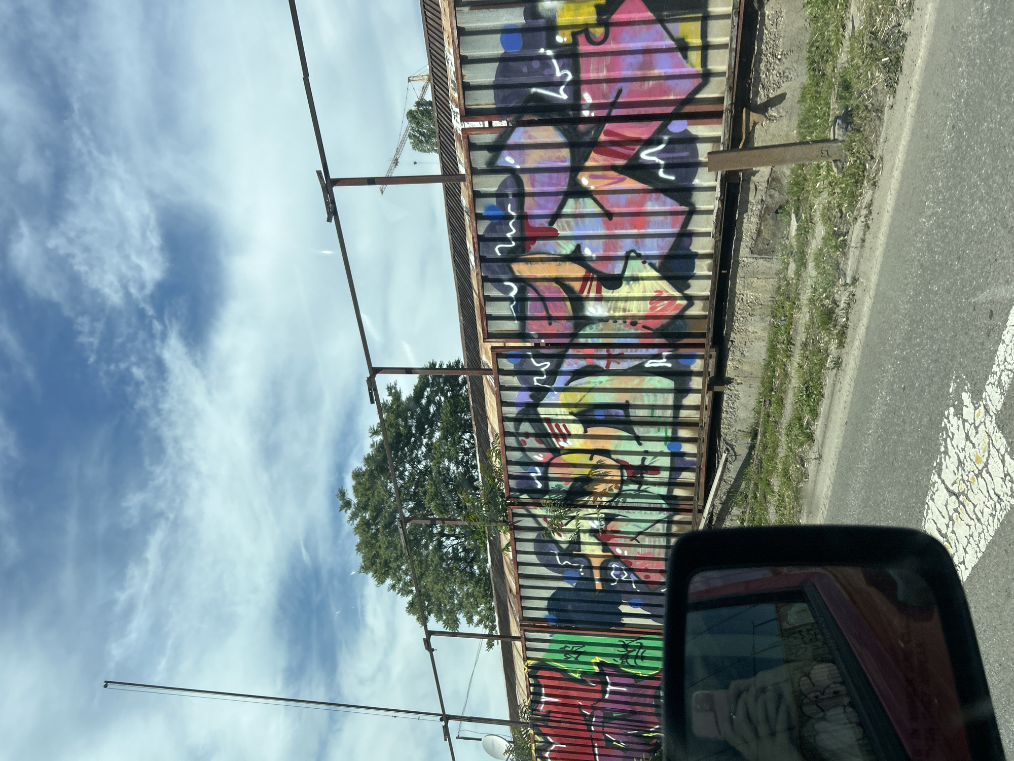 The image shows a fence covered in colorful graffiti, with a tree growing behind it. The fence appears to be constructed of metal corrugated panels. The background features a blue sky with scattered clouds. A crane is faintly visible in the distance. The image was taken from inside a vehicle, as evidenced by the car's side mirror in the foreground reflecting a hand holding a smartphone. The location is in the city of Sofia, Bulgaria.