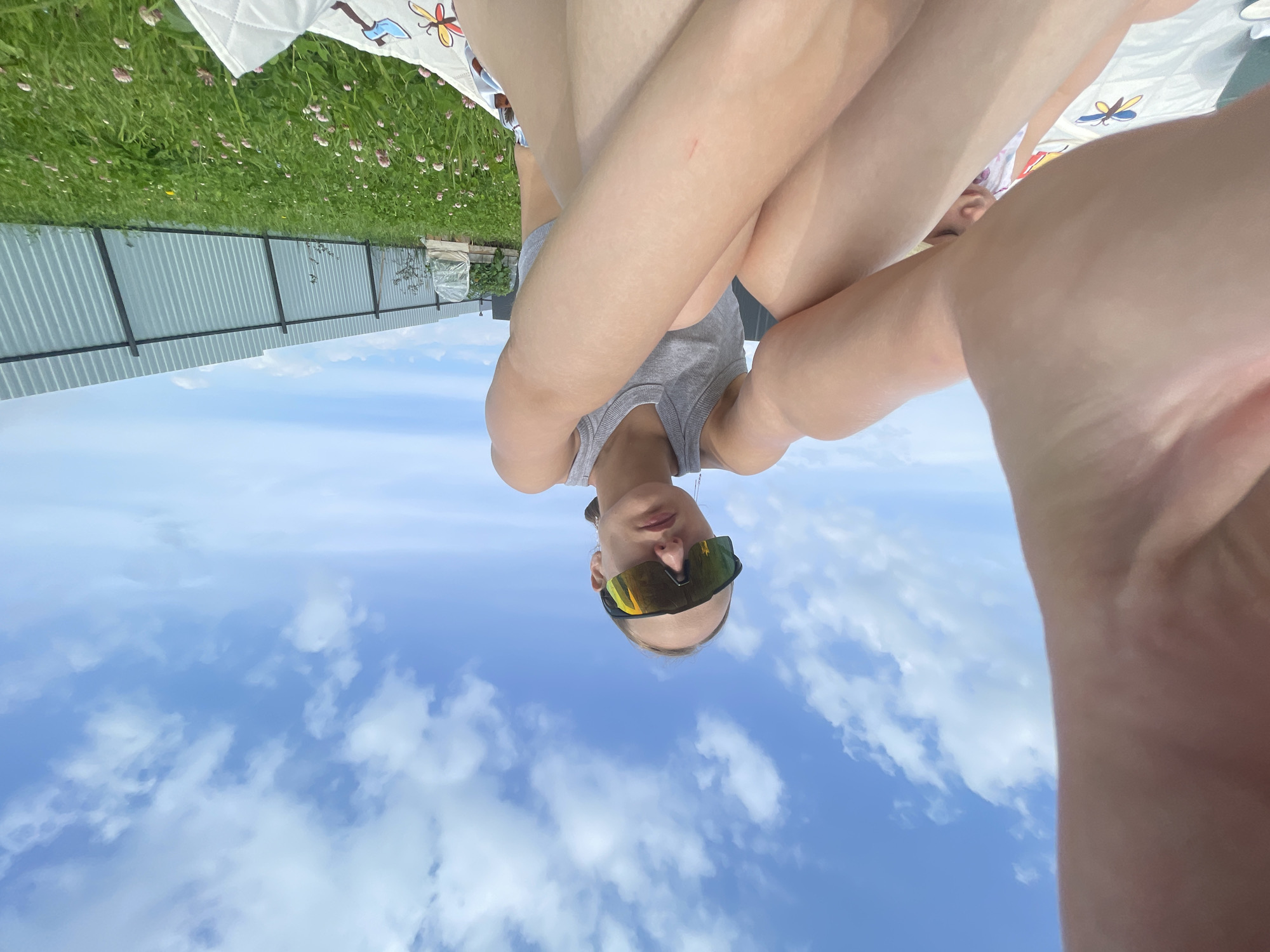 In the city of Podshivalovo, Russia, a young woman with blonde hair tied back, wearing large, reflective sunglasses and a gray tank top, sits on a white blanket spread on a green lawn. She is positioned outdoors, likely in a backyard, as evidenced by the presence of a metal fence in the background. The sky above is a vibrant blue with scattered white clouds. The woman is holding up her left arm, partially obscuring the view of a young child seated behind her, who is wearing a patterned garment. The overall scene depicts a relaxed outdoor activity.