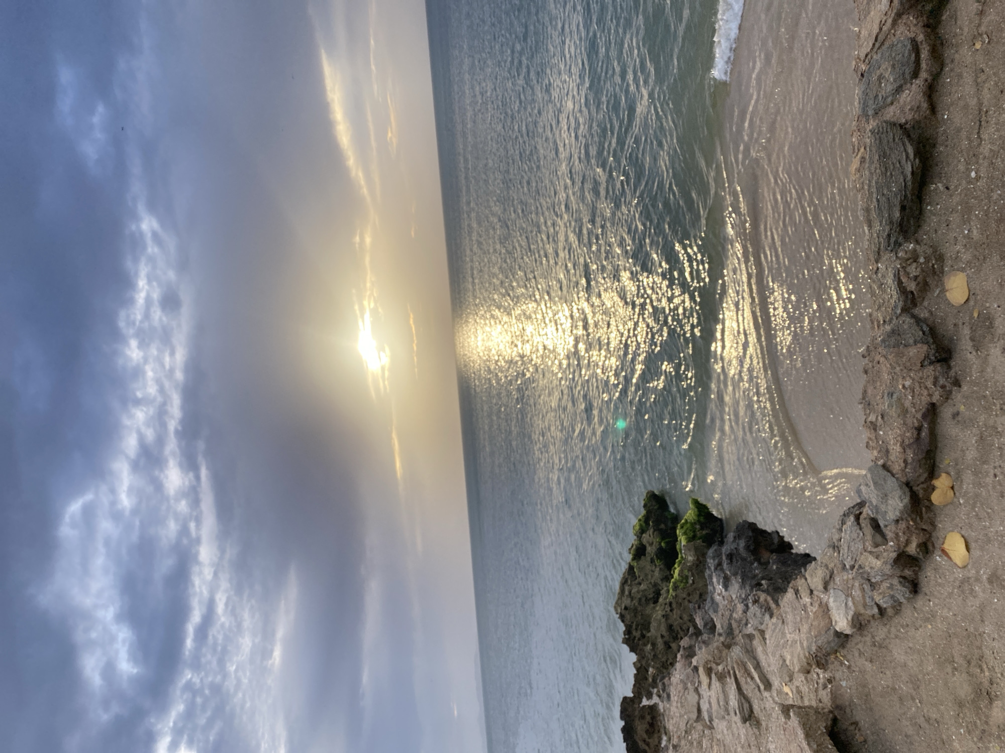 The image shows a natural, coastal landscape in Juan Griego, Venezuela, during sunset. The foreground features a rocky shoreline with a sandy embankment adorned with two fallen leaves. The water is calm and reflects the golden sunlight, creating a shimmering effect. A gentle wave breaks on the shore. The background consists of a vast ocean under a cloudy sky, with the sun partially obscured by clouds, casting rays of light. The horizon is visible in the distance, with a faint outline of land. No people or specific man-made structures are visible in the scene.