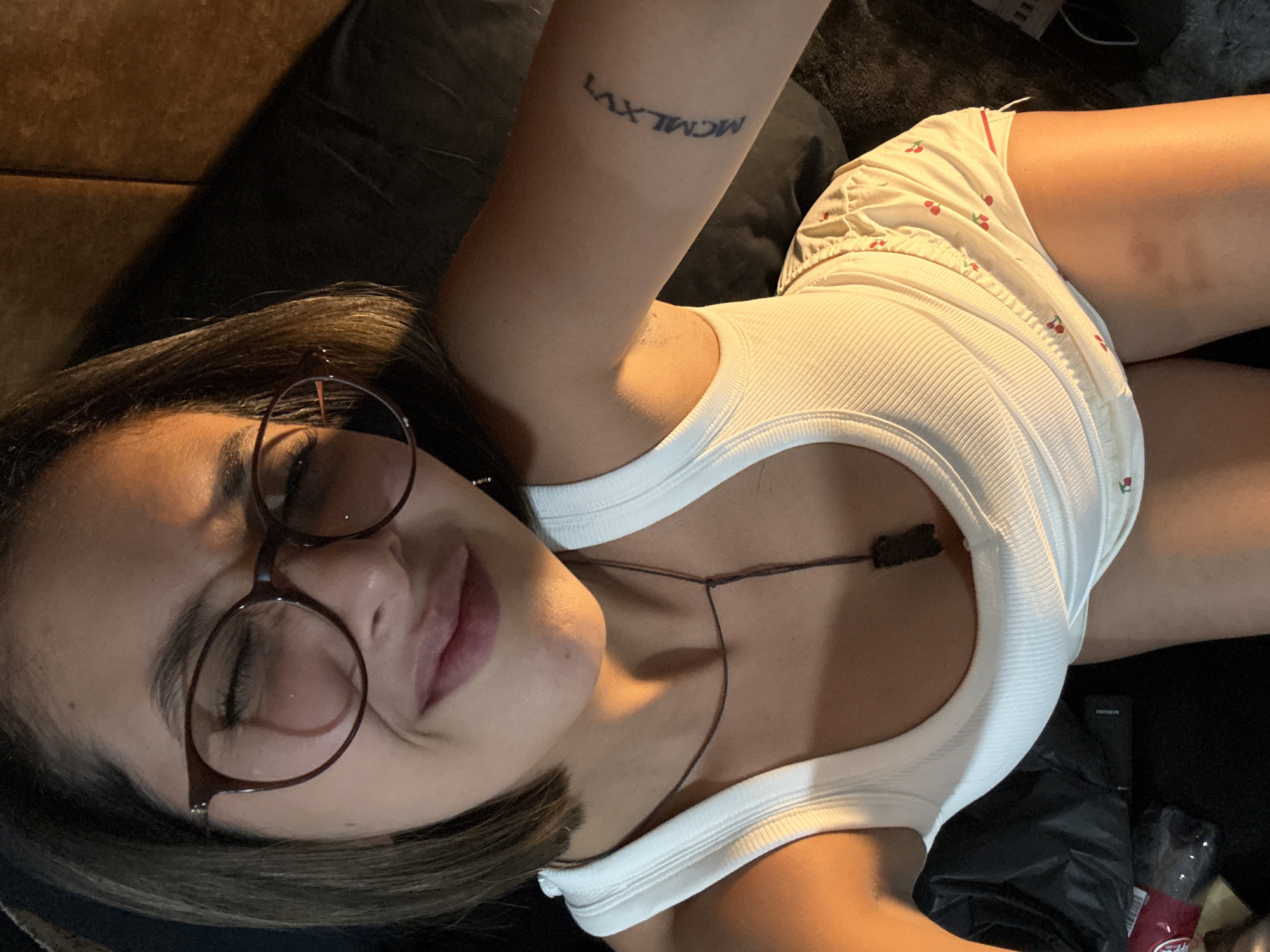 A woman wearing round eyeglasses and a white tank top is lying down. She has a tattoo on her left arm. She is wearing shorts with a cherry pattern. A water bottle is on the right side of the image. The location is not discernible from the image.