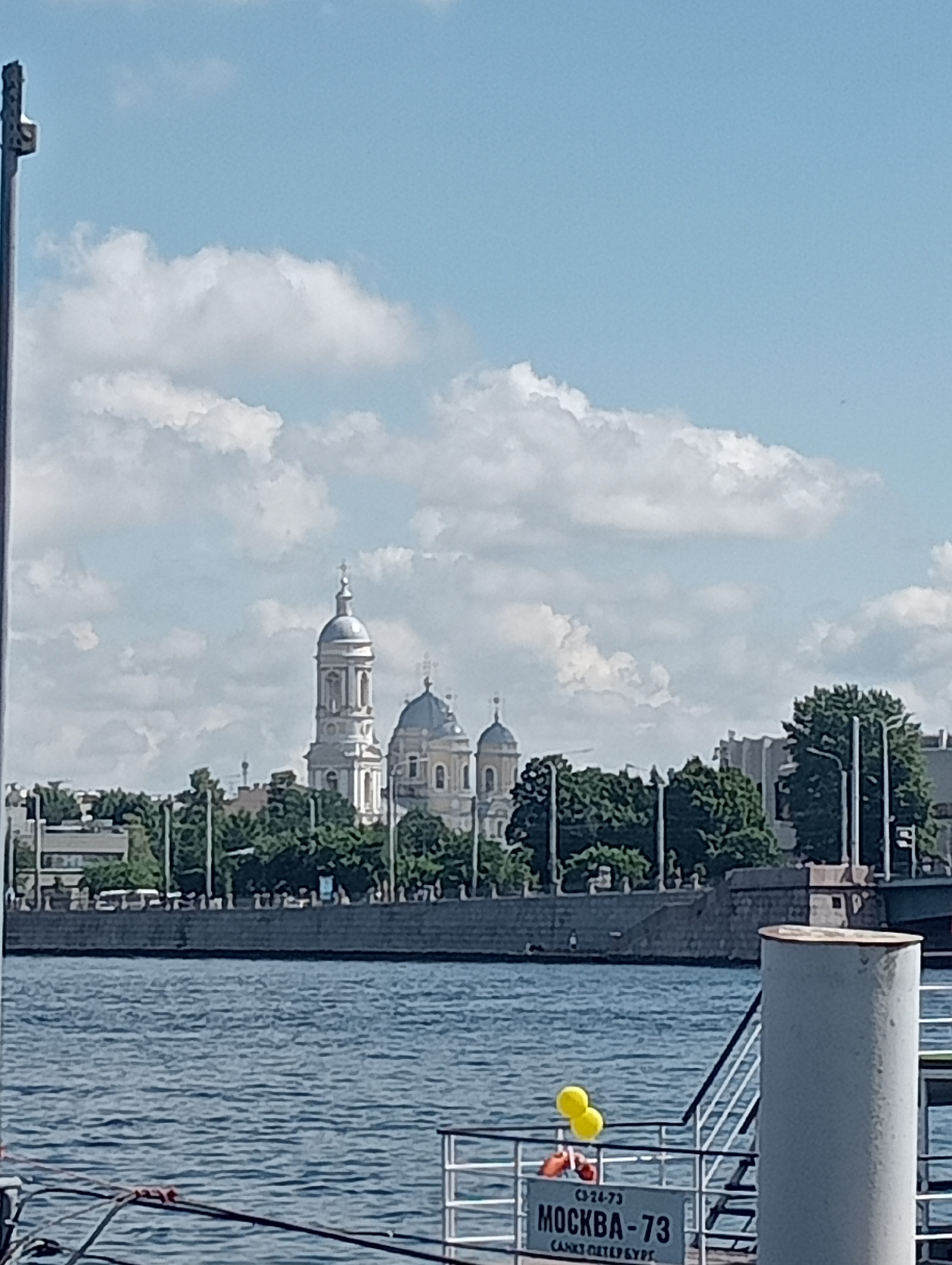 The image shows a view of the Smolny Cathedral in Saint Petersburg, Russia, across the Neva River. The cathedral's bell tower and domes are visible above a line of trees and buildings. In the foreground, a boat is docked, with a sign that reads "МОСКВА - 73". Two yellow balloons are tied to the railing of the boat. The sky is blue with scattered clouds. The overall scene depicts a sunny day on the river in Saint Petersburg.