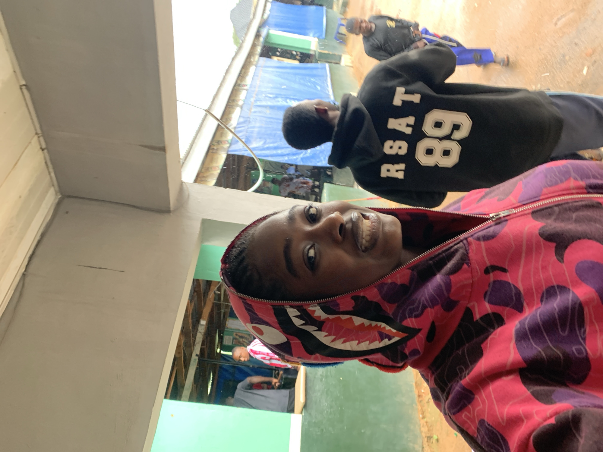 In New Stadium, Nigeria, a young woman with braided hair, wearing a pink and purple camouflage-patterned hoodie with a shark face graphic on the hood, smiles at the camera. Behind her, a young man in a black hoodie with "RSAT 89" printed on the back stands facing away. Further in the background, other individuals, including a man in a striped shirt and a person in a black long-sleeved shirt and blue pants, are visible. The scene appears to be an outdoor covered area, possibly a market or event space, with green walls and a corrugated metal roof. Blue tarps are visible in the background. The ground is a reddish-brown dirt.
