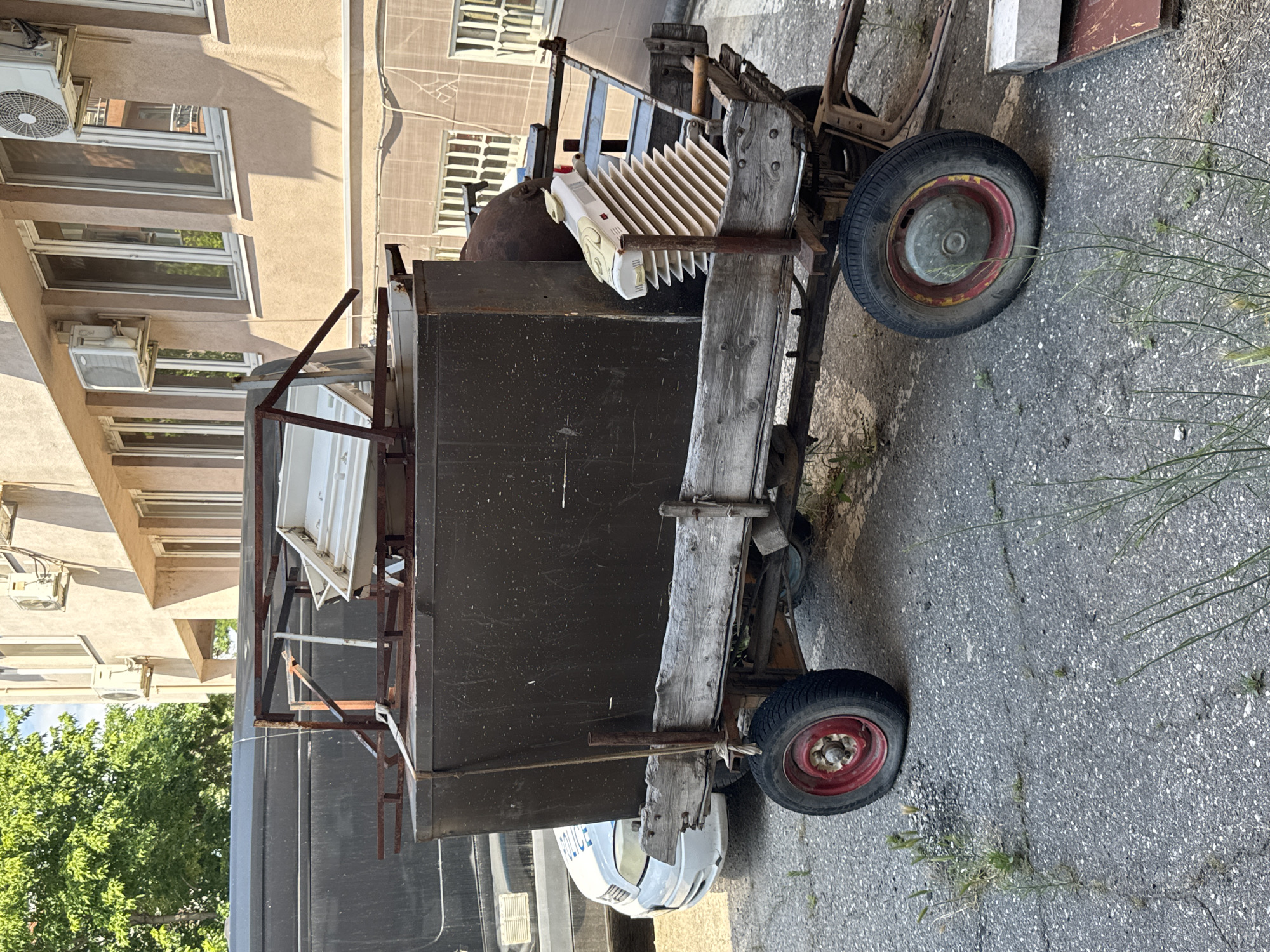 A large, weathered hand-pulled cart laden with various objects sits on a paved surface alongside a multi-story building. The cart features two wheels, each with a red center and a black tire. The frame is constructed from weathered wood and dark metal. On top of the cart, there's a mix of items including a white object with ridged texture, a dark metal container, and other indeterminate objects. A white vehicle with the word "POLICE" visible in reverse is parked behind the cart. The surrounding area is an urban environment with buildings and some vegetation. The location appears to be in a city.