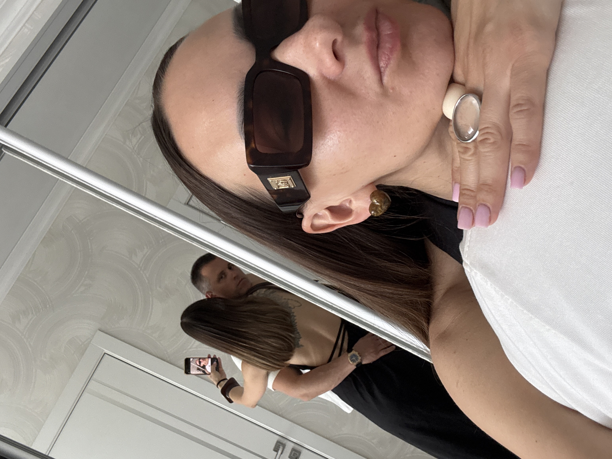 The image captures a mirror selfie taken indoors in what is identified as Sochi, Russia.
In the immediate foreground, a woman with long, dark hair is depicted from the upper chest upwards, with her right hand resting under her chin. She wears large, rectangular, tortoiseshell-patterned sunglasses featuring a gold emblem on the temple, and heart-shaped amber earrings. Her fingernails are painted light pink, and she wears a prominent, clear, oval-shaped ring on her right ring finger. She is dressed in a white top with a visible black strap on her left shoulder.
The reflection in the mirror reveals a second woman and a man embracing. The second woman, with long, dark hair, is shown from the back, wearing a black backless garment with thin straps crossing her lower back. A large, dark tattoo of a winged creature is visible on her upper back. She holds a smartphone in her right hand, angled to take a photo, with a partial reflection of a face visible on its screen. The man, standing behind her with his right arm around her waist, wears a white short-sleeved t-shirt and a dark watch on his left wrist. He has short, dark hair and looks directly towards the mirror with a neutral expression.
The background shows a light-colored wall with a subtle, circular or wavy patterned wallpaper. A white door with a modern rectangular handle is visible on the left side of the reflection.