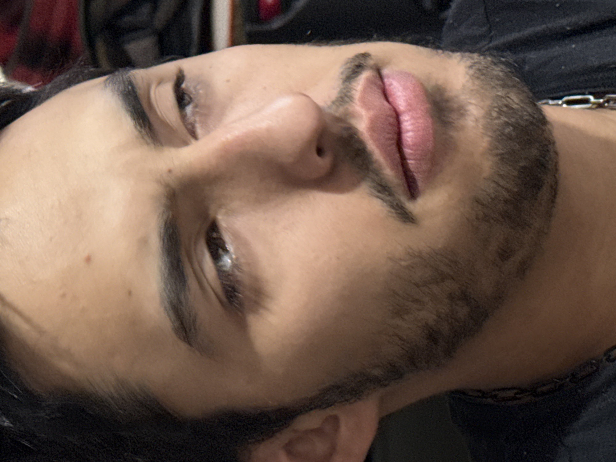A close-up shot shows a person with olive skin, dark brown hair, and a beard. The person is lying down, angled slightly upwards towards the camera. Their eyes are partially closed, and their expression appears relaxed. The person has full lips and a well-defined nose. A silver chain is visible around their neck, and they are wearing a dark shirt. The background is out of focus, but it seems to consist of various soft items. The photograph was most likely taken in an indoor setting, such as a bedroom or living room, somewhere in Puebla, Mexico.