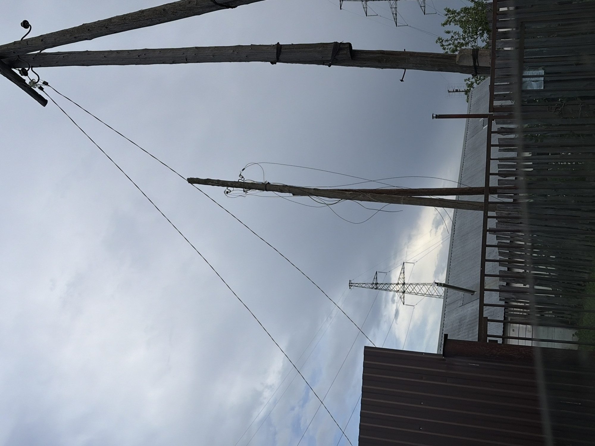 The image depicts a cloudy sky above a scene in what appears to be a residential or industrial area. Several wooden utility poles with wires extend across the frame. A metal fence is partially visible on the right. A corrugated metal structure is present in the foreground. In the background, a power transmission tower is visible. Based on the provided information, the image is located in the city of Chaikovsky, Russia.