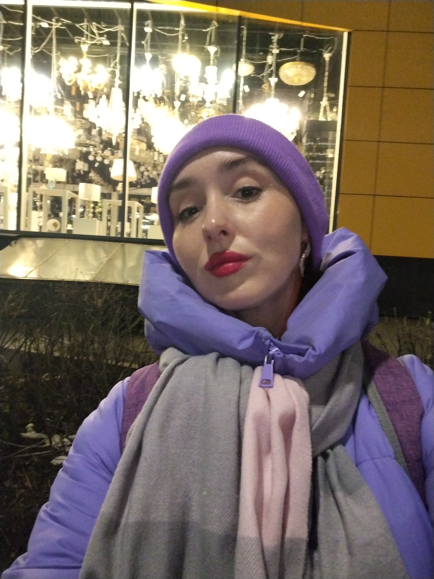 A person with fair skin and red lipstick is wearing a purple beanie, jacket and a gray and pink scarf. The person is standing outside at night, with a storefront window in the background displaying multiple chandeliers. The location is possibly Izhevsk, Russia. Bushes are in the foreground. The building exterior is yellow.