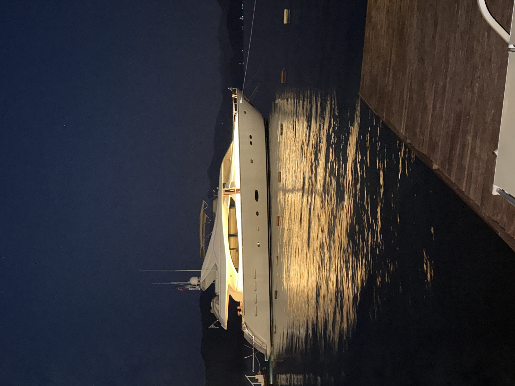 A large, white yacht is moored at night in the harbor of Marmaris, Turkey. The yacht is illuminated by interior lights, casting a warm glow on the water. The water is dark and still, reflecting the yacht's lights in shimmering patterns. In the foreground, a wooden dock juts out into the water. The sky is a deep navy blue, with a few faint stars visible. The silhouette of mountains can be seen in the distance. No people are visible on the yacht or the dock.