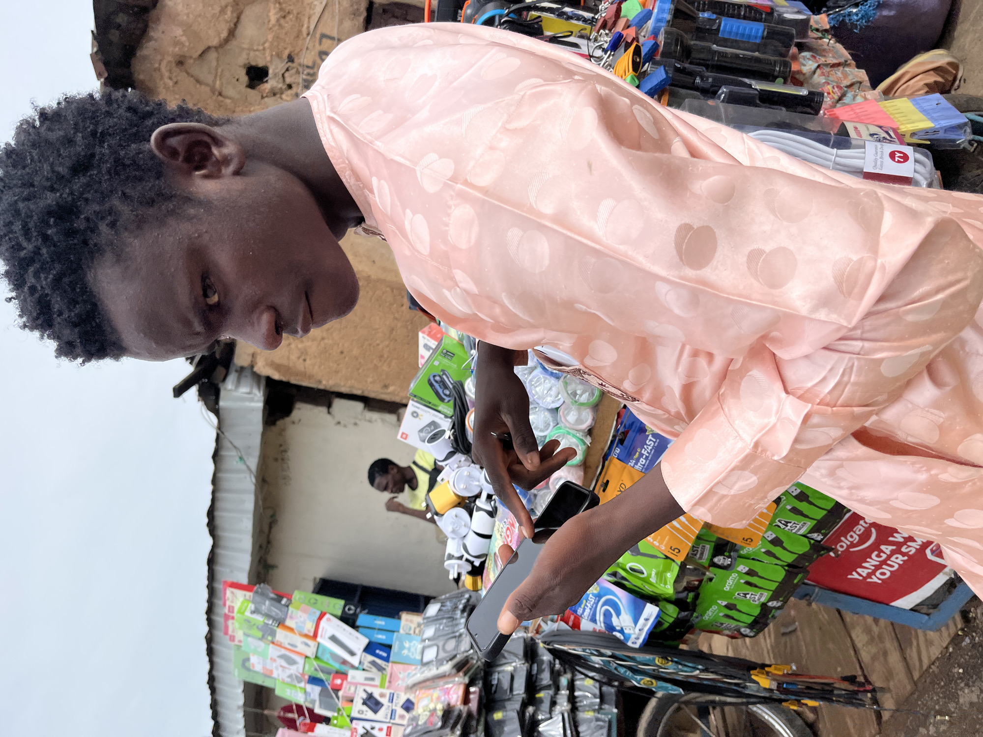 The image captures a vibrant market scene in Jos, Nigeria, focusing on a young Black man examining a smartphone at a stall filled with electronic goods. The main subject, positioned on the left side of the frame, is seen in profile, looking intently downwards at a dark-colored smartphone held in both hands. He has short, curly black hair and wears a light pink, possibly satin or silk, long-sleeved garment adorned with a subtle pattern of circles or hearts. His expression is serious and focused.
In the background, another individual wearing a yellow top is partially visible and blurred, suggesting a busy market environment. The market stall itself is densely packed with various merchandise, primarily electronic accessories. These include numerous packaged USB cables, fast chargers, power adapters, small flashlights, and other consumer electronics. The background features parts of a building structure and an overcast, bright sky, indicating daytime.
Visible text on the product packaging includes brand names and descriptive words such as "Cmnl-FAST," "FAST," "tra-FAST," "aramo," "oramo," and "Colgate." Other snippets read "YANGA W YOUR SM," "Brick De," and "7Y." The number "15" is also frequently seen on various packages, likely indicating a product specification or model. The scene conveys the bustling atmosphere of an outdoor market.