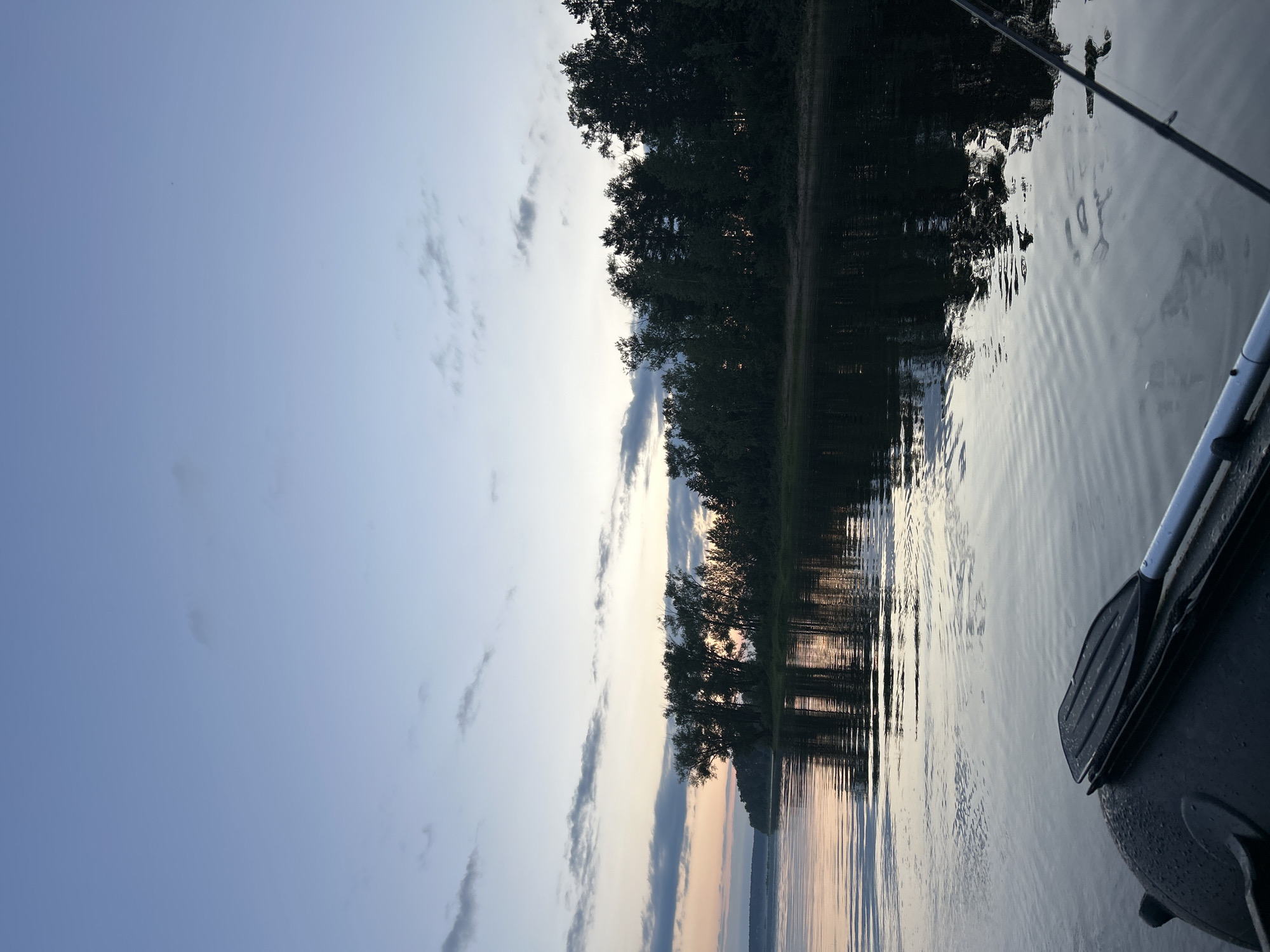 The image depicts a serene scene on a body of water, likely a lake or river, at dusk. The sky is a soft gradient of blues and grays, with scattered clouds tinged with the warm hues of sunset. Silhouetted against the sky is a dense line of trees along the shoreline, their reflections shimmering on the water's surface. The water itself is calm, with gentle ripples creating a textured pattern. In the foreground, a portion of a kayak or small boat is visible, with a paddle resting on its edge. The paddle appears to be made of plastic or a similar material, with a dark blade and a lighter shaft. A fishing rod is also partially visible, extending diagonally across the upper right portion of the frame. The overall mood is tranquil and peaceful, suggesting a moment of quiet contemplation or leisurely activity on the water. The time of day is clearly evening, as indicated by the soft light and the color of the sky. There is no visible text in the image. The setting evokes a sense of natural beauty and solitude.