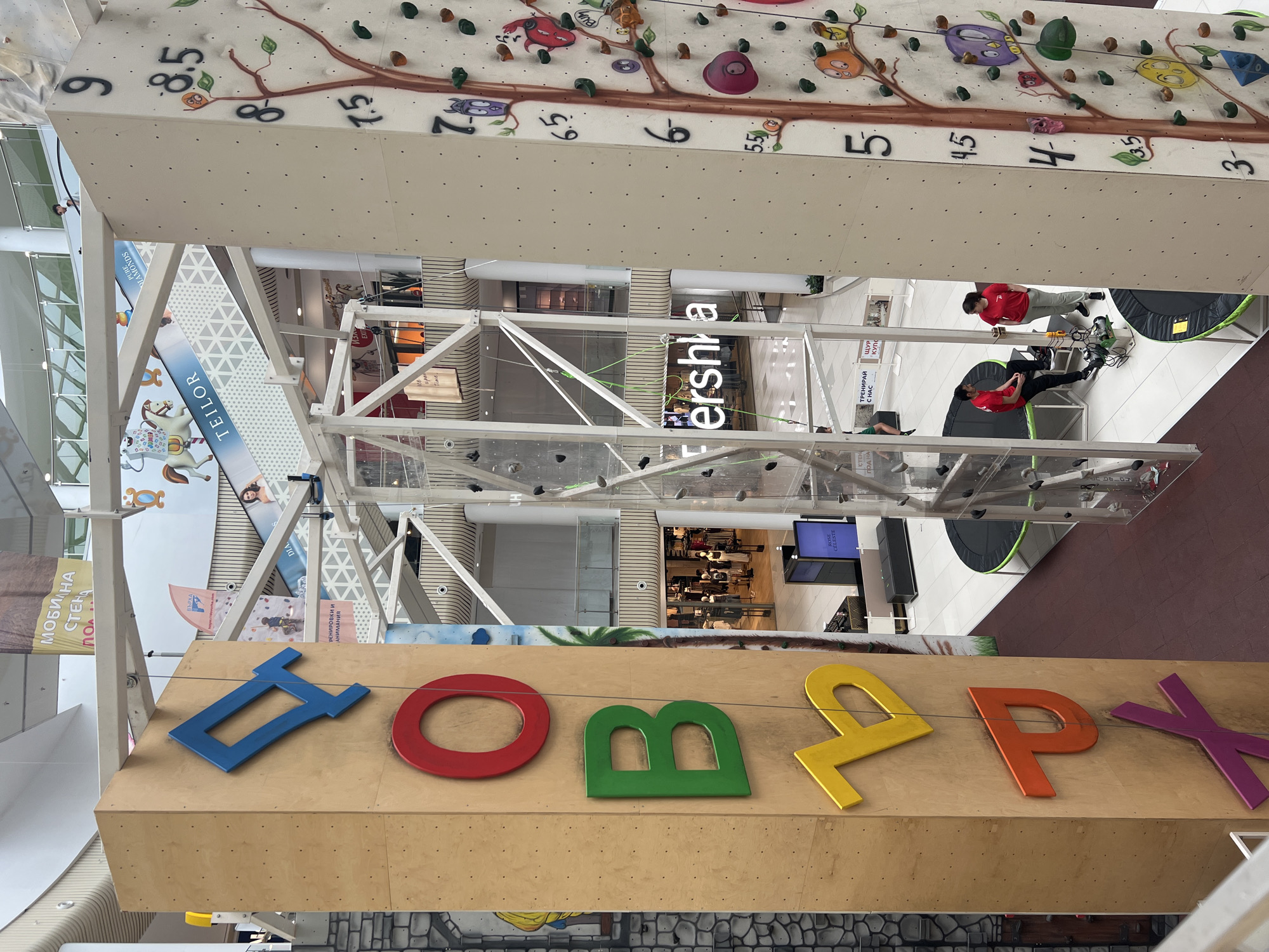 This image captures a vibrant indoor climbing facility, likely situated within a multi-story shopping mall in Plovdiv, Bulgaria. The primary subject is a large, light-colored climbing wall, adorned with colorful holds, painted tree branches, leaves, and whimsical cartoon creatures, creating a playful and inviting atmosphere. Numbers such as "9", "8.5", "6", "5.5", "5", "4.5", "4", "3.5", and "3" are painted on the wall, likely indicating route difficulty or height markers.
In the foreground, a wooden structure displays large, colorful three-dimensional Cyrillic letters spelling "ДОБЪРХ" (DOBЪRH), possibly a brand name or a playful slogan. Below the climbing wall, the multi-level architecture of the mall is visible, featuring glass barriers and various storefronts.
Several people are present. Two individuals wearing red shirts appear to be directly involved with the climbing activity, one standing beside climbing gear like ropes and harnesses, and another sitting on a mat. A person in a green shirt is also visible near the climbing structure. Other individuals are seen on the lower mall levels, presumably shoppers or visitors.
Visible text on banners and signs within the mall includes "МОБИЛНА СТЕНА" (Mobile Wall), "ВЪРХА" (The Peak), "ТРЕНИРАЙ С НАС" (Train with Us), and "ЩУР КУПОН" (Crazy Party), all related to the climbing activity. Commercial signage for "Bershka", "TEILOR", and "PURE DIAMONDS" is also clearly visible, reinforcing the shopping mall setting. The scene depicts a lively, recreational environment, well-lit as if during daytime.