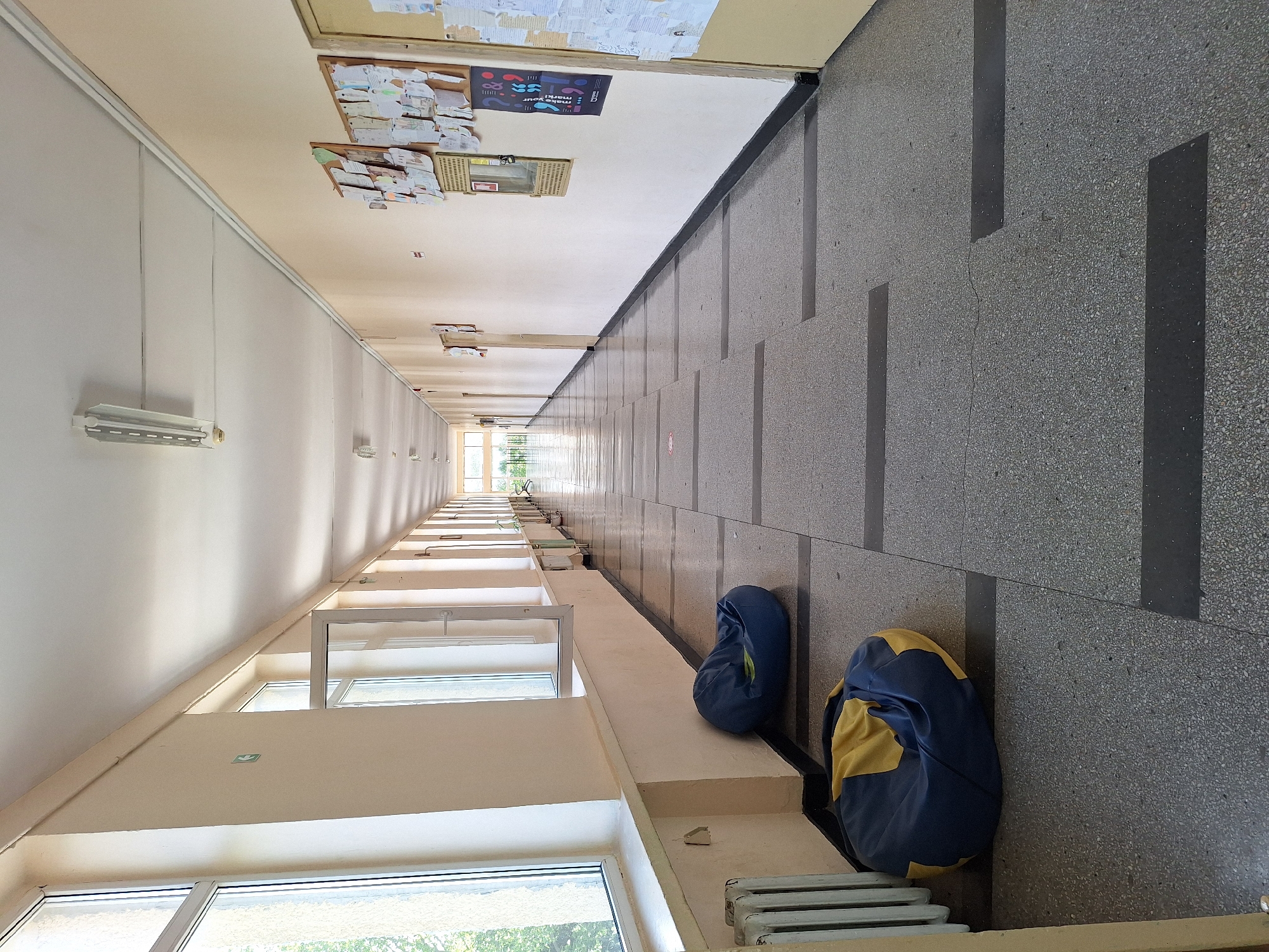 The image depicts a long, empty school hallway in Varna, Bulgaria, on a bright day. The hallway has a tiled floor with dark gray rectangular inserts. Along the left wall, there are several large windows and a white radiator. Two beanbag chairs, one blue and yellow and the other entirely blue, are placed on the floor near the windows. The right wall is painted a light beige and features bulletin boards with various papers and posters, as well as several doors. Fluorescent light fixtures are mounted on the white ceiling. The hallway extends into the distance, creating a sense of depth.