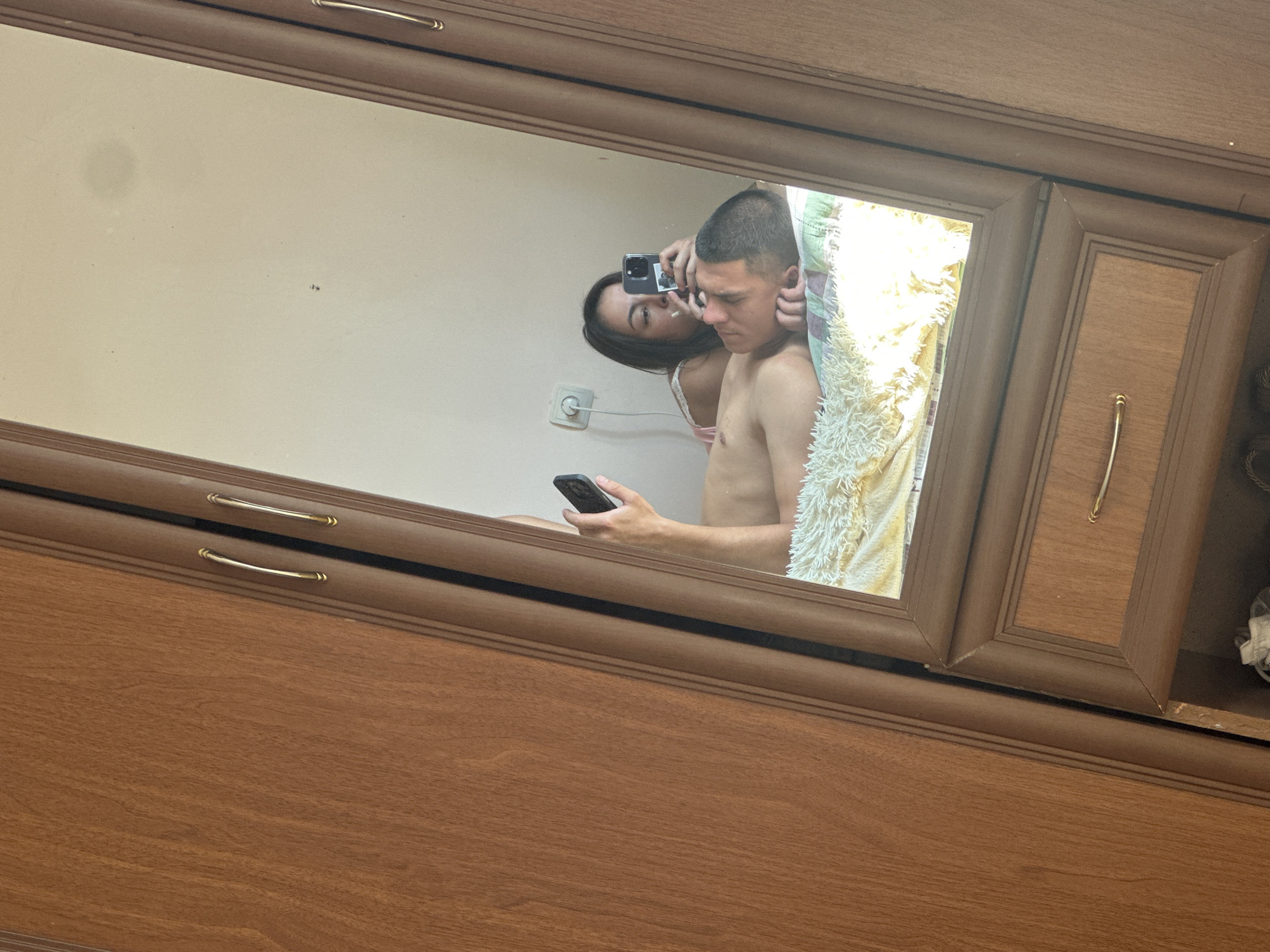 A mirror in a wooden frame reflects a couple lying on a bed. The man is lying on his back, shirtless, with his head resting on a pillow. The woman is lying on her side, leaning on her elbow, and looking at her phone. She is holding another phone in her hand and appears to be taking a picture or video of herself and the man. The bed is covered with a yellow and white patterned blanket. In the background, a white wall with a light switch and a power outlet is visible. The image is taken from an angle that shows a wooden wardrobe with two doors and gold handles on the left side of the frame. The setting appears to be a bedroom.