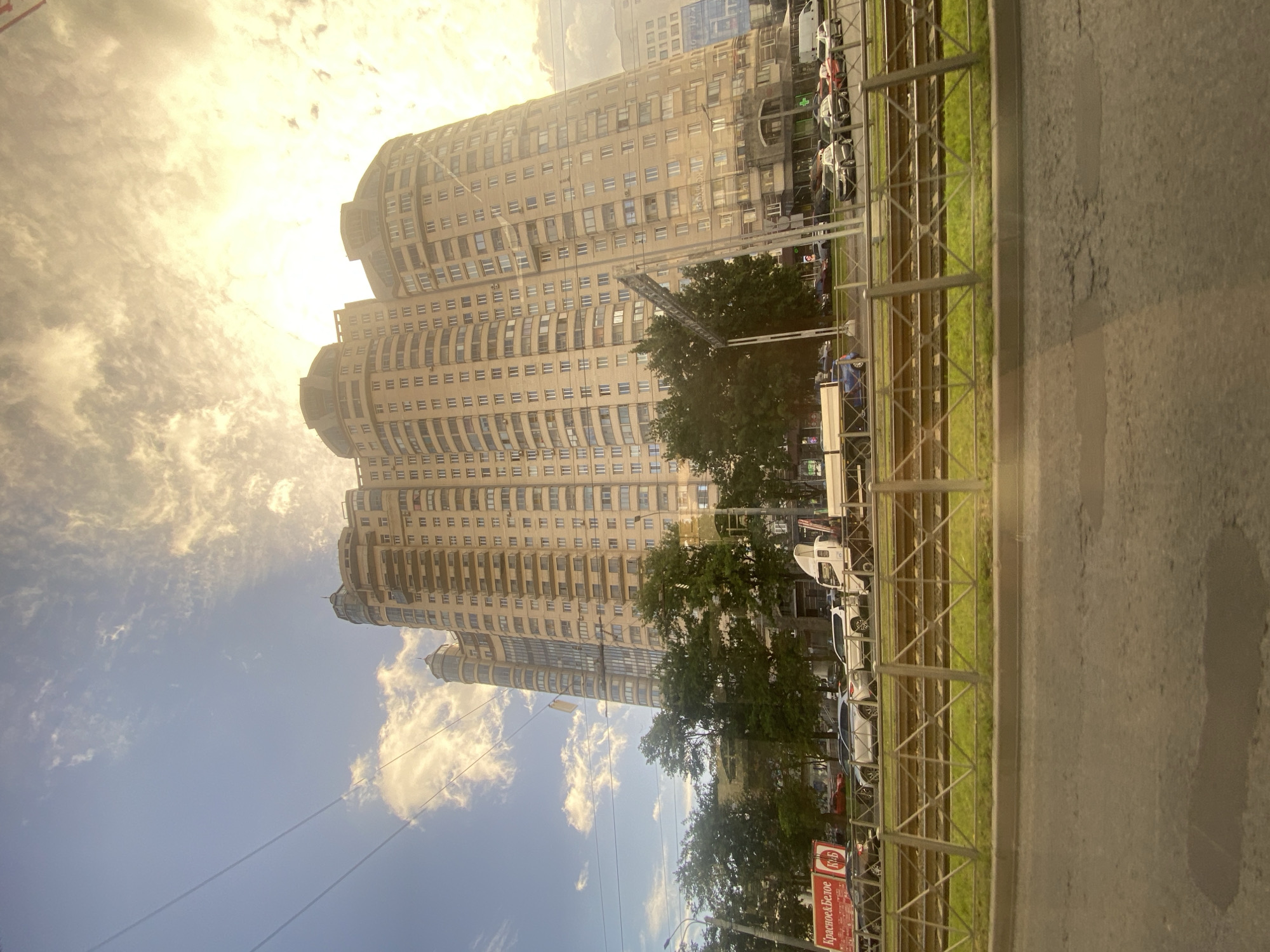 The image captures a large, modern, light-beige residential building with a curved facade, dominating the upper right portion of the frame. The building features numerous windows and balconies, and its top is partially obscured by a bright, cloudy sky illuminated by sunlight, creating a lens flare effect. In the foreground, a busy street scene unfolds with several cars parked and driving. There are also tram tracks visible behind a metal fence. Green trees line the street between the buildings and the road. A red and white sign with Russian text, "Красное & Белое," is visible on the left side, indicating a store. The overall impression is of an urban environment in Saint Petersburg, Russia, during daylight hours, likely late afternoon due to the warm lighting.