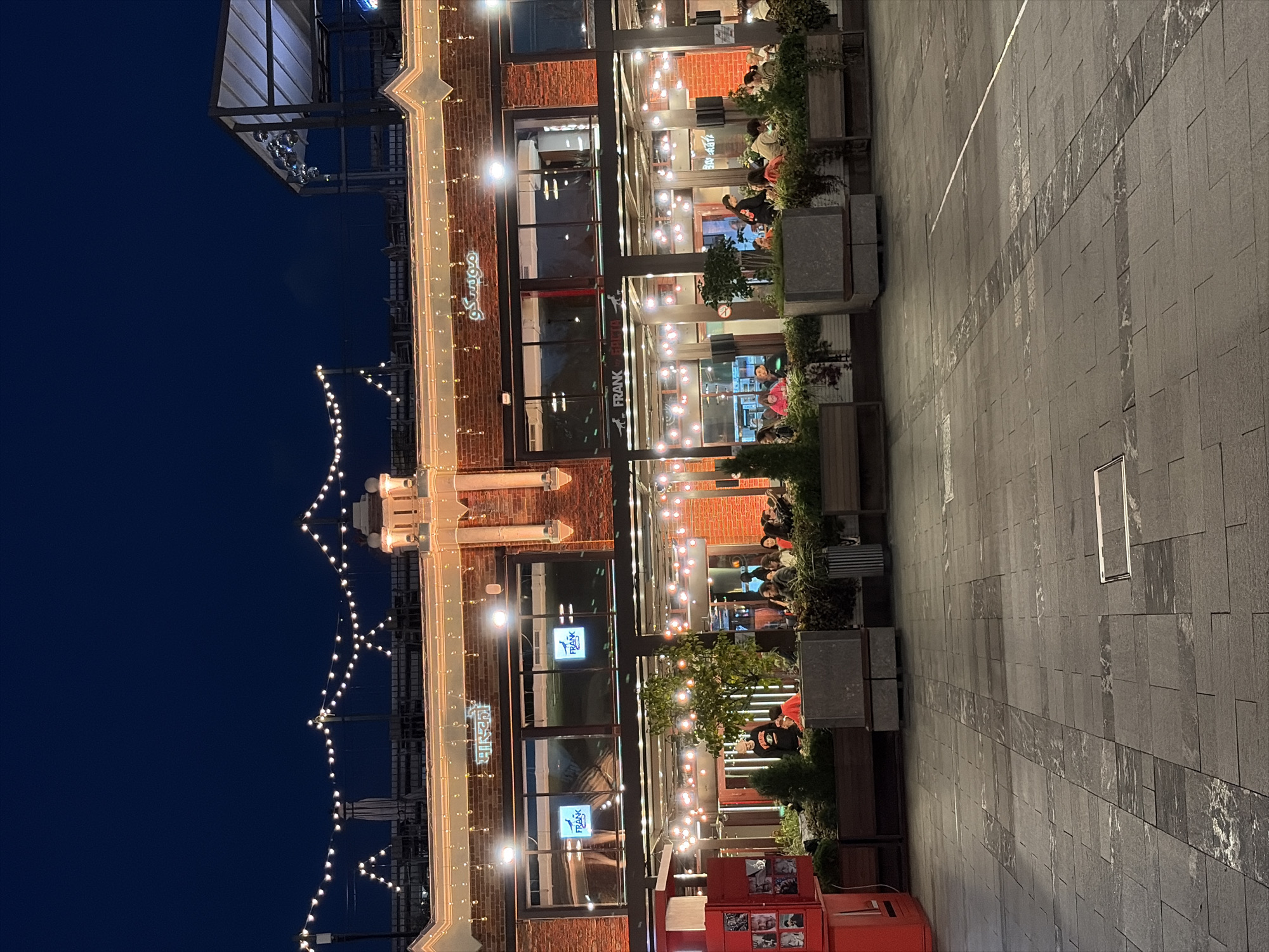 The image depicts an outdoor dining area of a restaurant complex at dusk, likely in Moscow, Russia, given the signage which includes Russian text. The building facade is constructed of red brick and is illuminated by decorative string lights and internal lighting, creating a warm ambiance. Above the storefronts, a sign with Cyrillic lettering reads "МУСЛИН КО" (Muslin Co.), and another sign in Hindi characters appears to say "मास्क" (Mask). Several windows on the upper level display illuminated logos for "FRANK".
Below the upper level, a covered patio area is furnished with tables and chairs, occupied by patrons engaged in dining and conversation. Lush planters filled with greenery and trees are strategically placed throughout the outdoor seating area, softening the urban environment. In the foreground, a paved pedestrian walkway is laid with large, dark grey tiles, featuring a decorative inlay pattern. To the left, a bright red vending machine or information kiosk is visible. The overall scene suggests a lively evening atmosphere at a popular dining establishment.