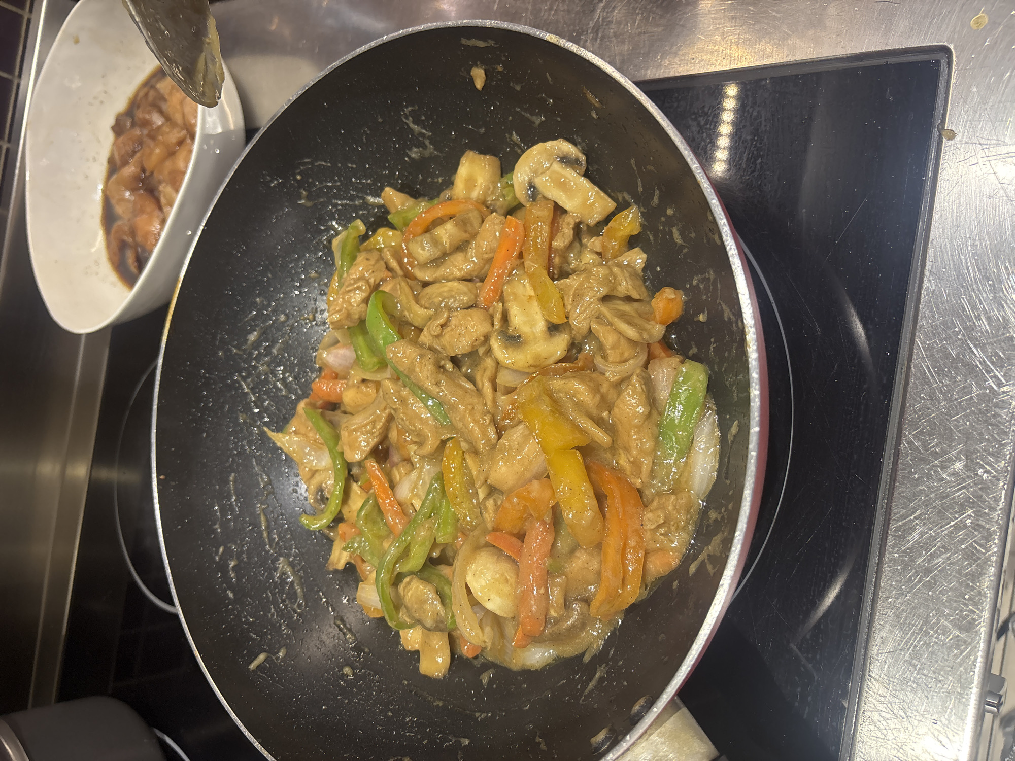 A wok full of stir-fried chicken and vegetables sits on a stovetop. The dish contains sliced chicken, bell peppers of various colors, mushrooms, and onions coated in a light brown sauce. In the background, a white bowl holds raw chicken pieces marinating in a dark liquid. The scene appears to be in a kitchen, likely in Dubai, United Arab Emirates, based on the context provided. The stovetop is modern and black, and the surrounding counters are metallic.