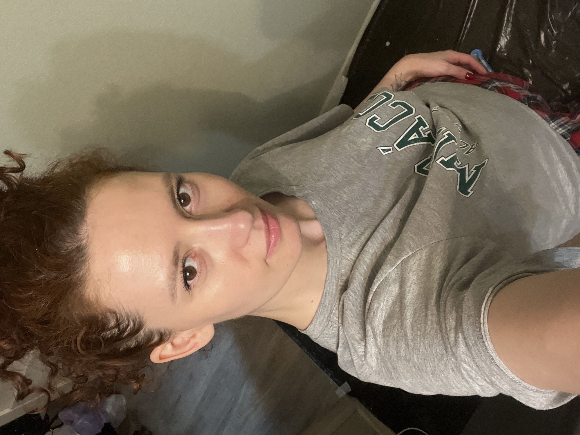 An indoor, high-angle selfie captures a female individual in Saint Petersburg, Russia. The subject, displaying fair skin, reddish-brown curly hair styled in a bun, and brown eyes with minimal makeup, gazes directly into the camera. She wears a light grey short-sleeved t-shirt featuring a partial green and white graphic (reading "MÁC" or "MACU") and red-and-blue plaid bottoms. Her left hand, with red painted fingernails and a small, faded tattoo on the inner forearm, rests gently on a noticeably rounded abdomen. The background consists of a muted sage-green wall on the right and a dark floor, potentially covered with a dark sheet or tarp, in the lower right. Indistinguishable objects are partially visible in the blurry upper left background.