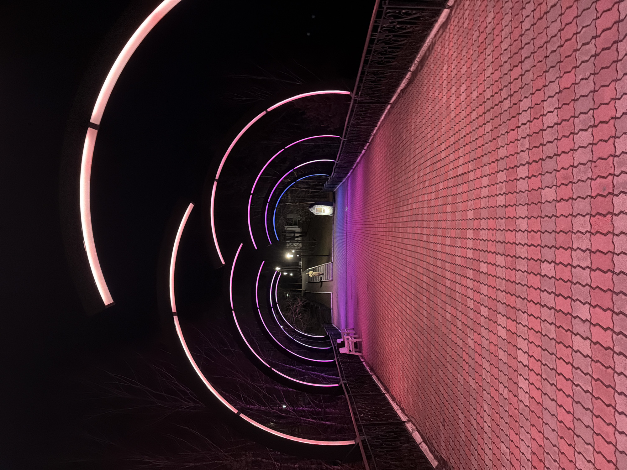 The image shows a pedestrian walkway in Слизнево, Russia, illuminated by a series of curved, colored light installations. The walkway is paved with interlocking red and gray stones. Metal railings run along the side of the walkway. The lighting transitions from pink to purple to blue as it recedes into the distance. The background is dark, suggesting it is nighttime. Bare tree branches are visible near the light installations. There are other lights further down the path. A bench is visible on the right side.