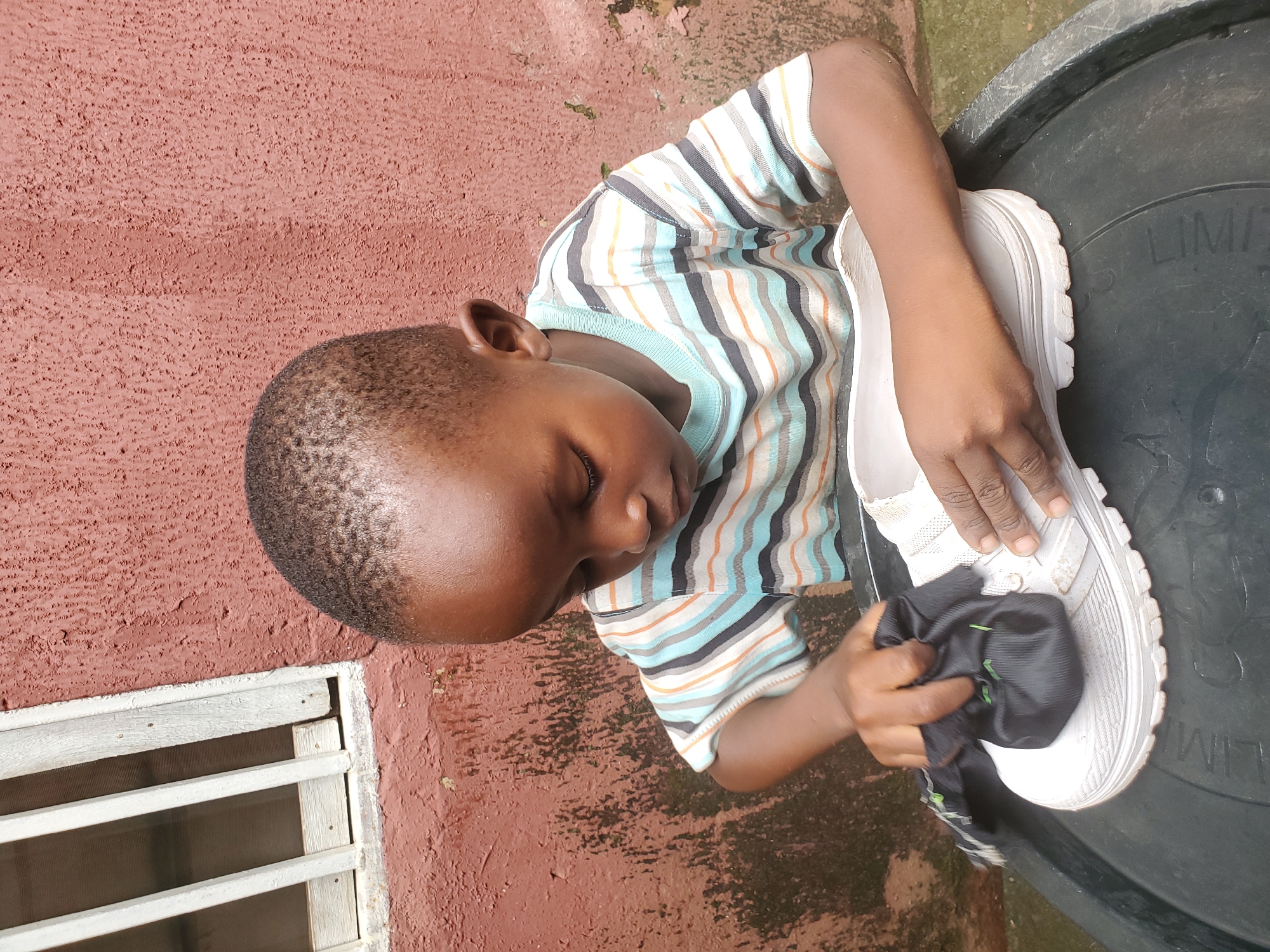 A young boy in a striped t-shirt is cleaning a white shoe with a black cloth. He is sitting next to a large, dark container, possibly a water tank, outdoors. The background is a textured, reddish-brown wall with some green patches, possibly moss or algae. To the left of the boy, a window with horizontal white bars is visible. The overall lighting suggests it is daytime. The boy appears focused on his task. The word "LIMIT" is visible on the dark container.